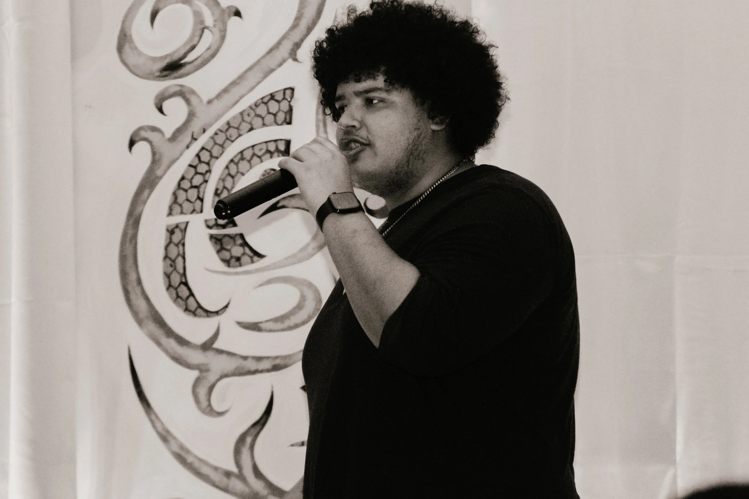 A young man with curly hair holding a microphone and singing or speaking, standing in front of a decorative wall with an intricate pattern.