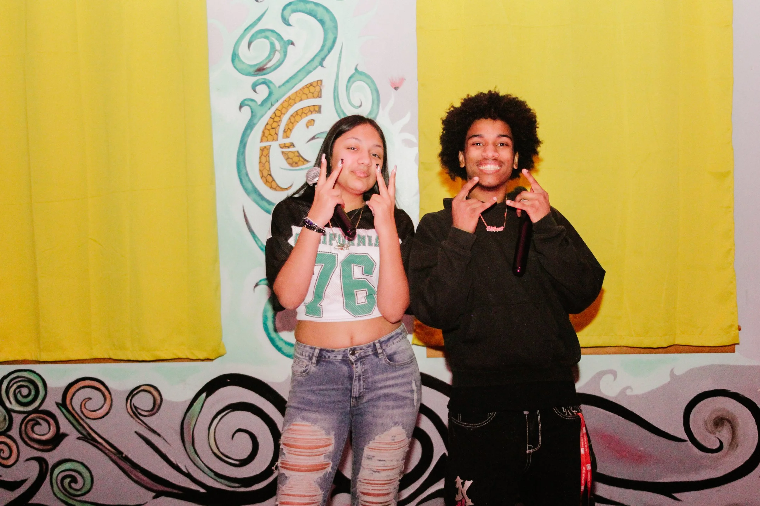 Two young people posing in front of a colorful mural, making peace signs and smiling. The background includes yellow curtains and a swirly black and white design.