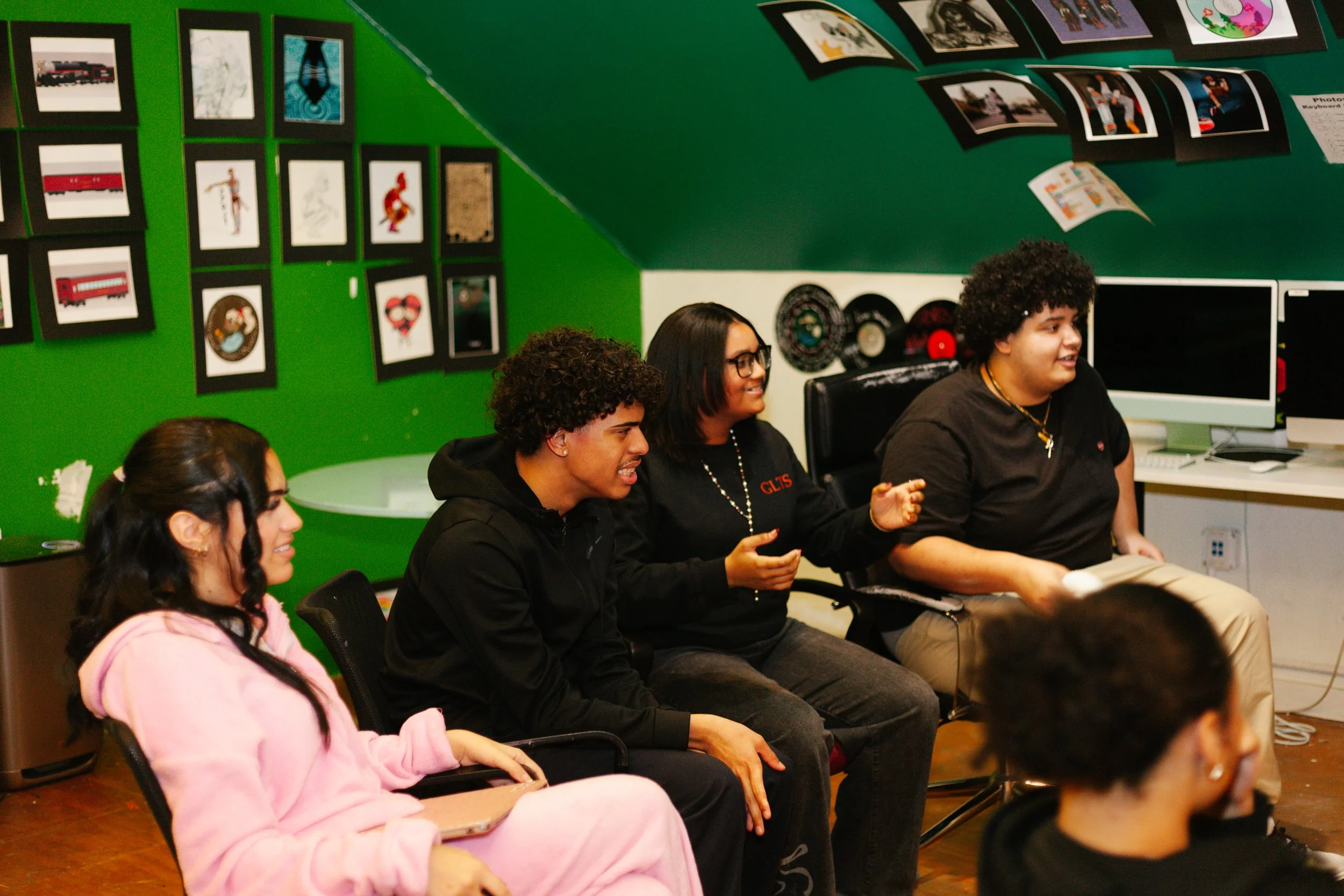 Group of diverse young adults sitting in a room, engaging in a discussion or activity, with green walls decorated with framed pictures and posters, and computers on desks in the background.
