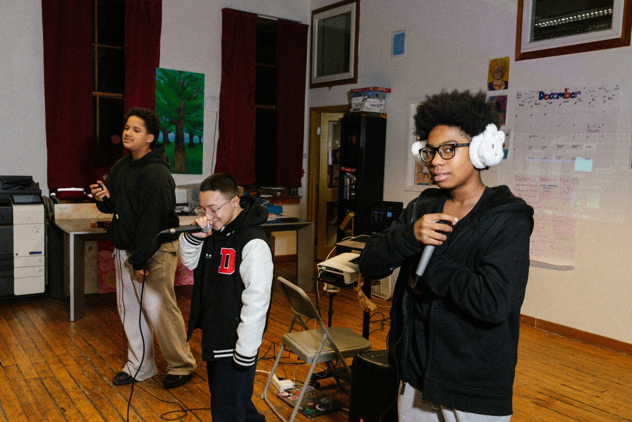 Three children singing into microphones in a classroom with a whiteboard and colorful posters.