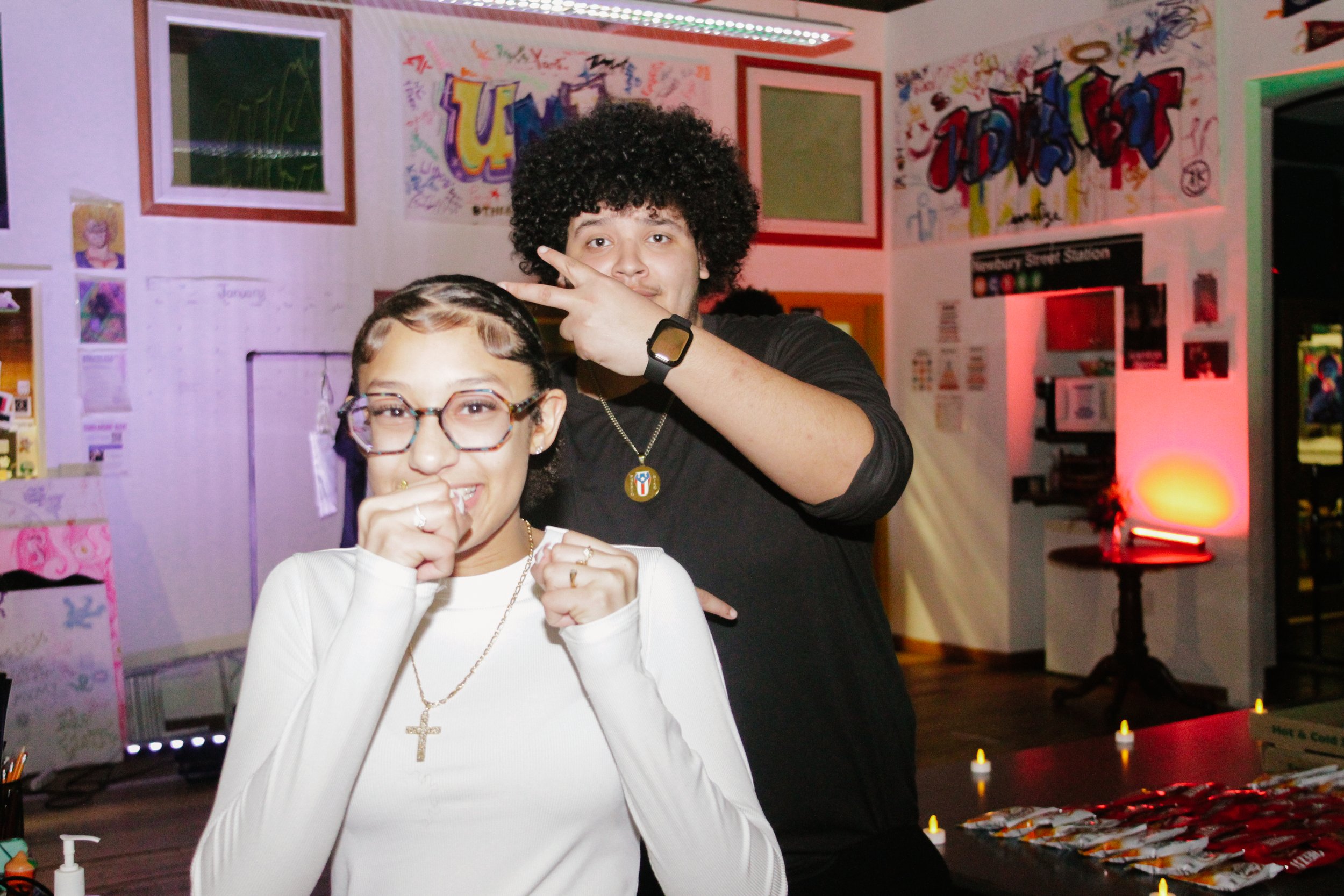 A girl with glasses and a girl with curly hair making peace signs at a colorful party or event.