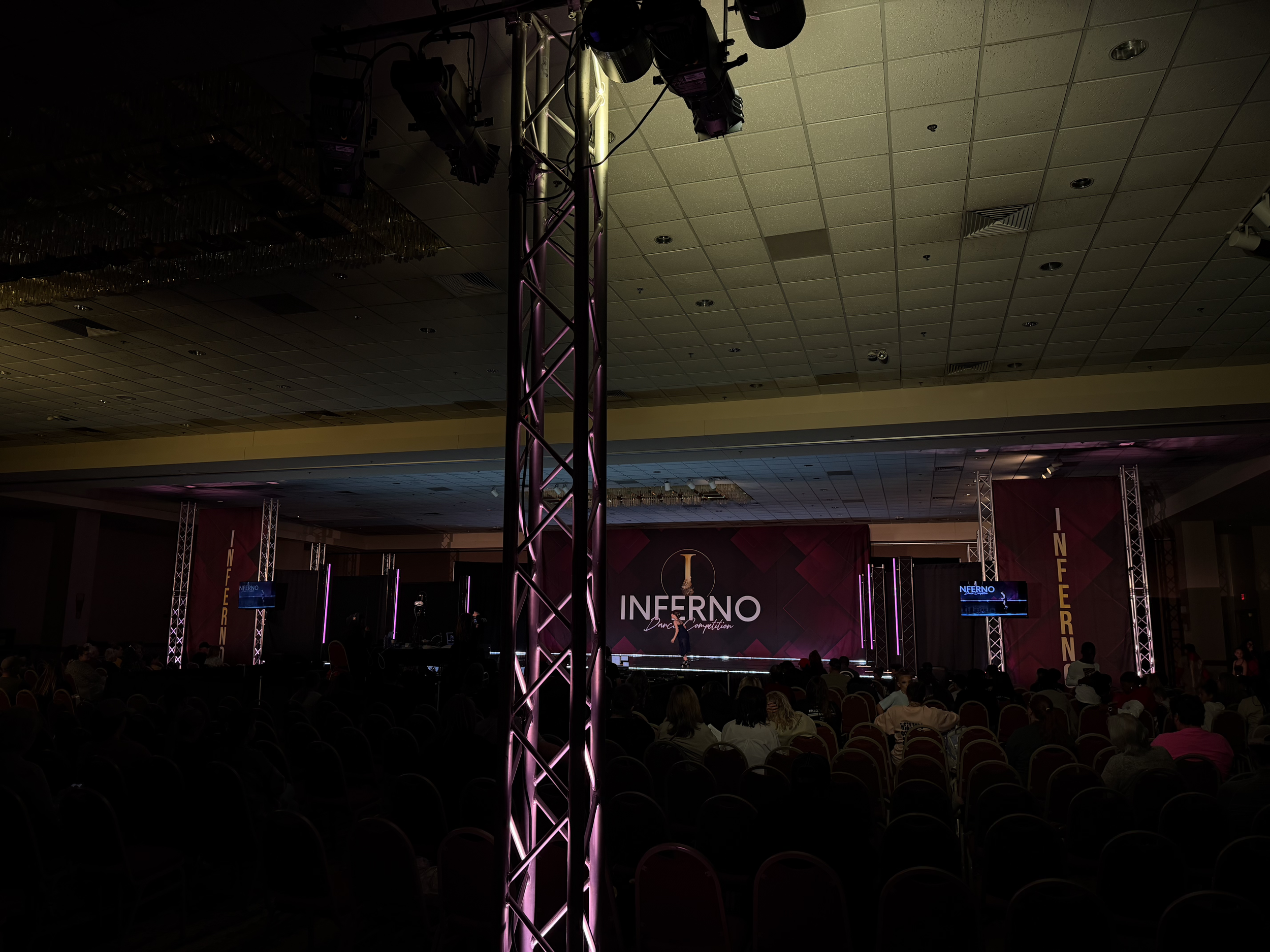 An indoor dance competition stage with purple lighting and a large screen displaying 'INFERNO Dance Competition'. Rows of chairs and audience members are visible in the dimly lit foreground.