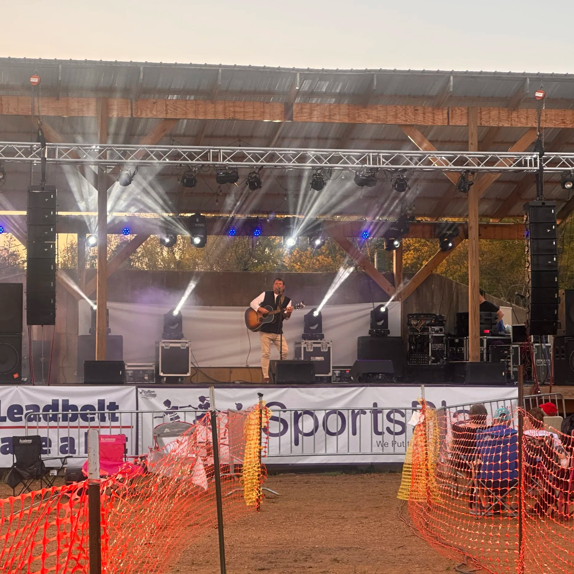 A band performs on an outdoor stage with wooden beams and a roof, bright stage lights and fog, and a man playing an acoustic guitar and singing into a microphone.