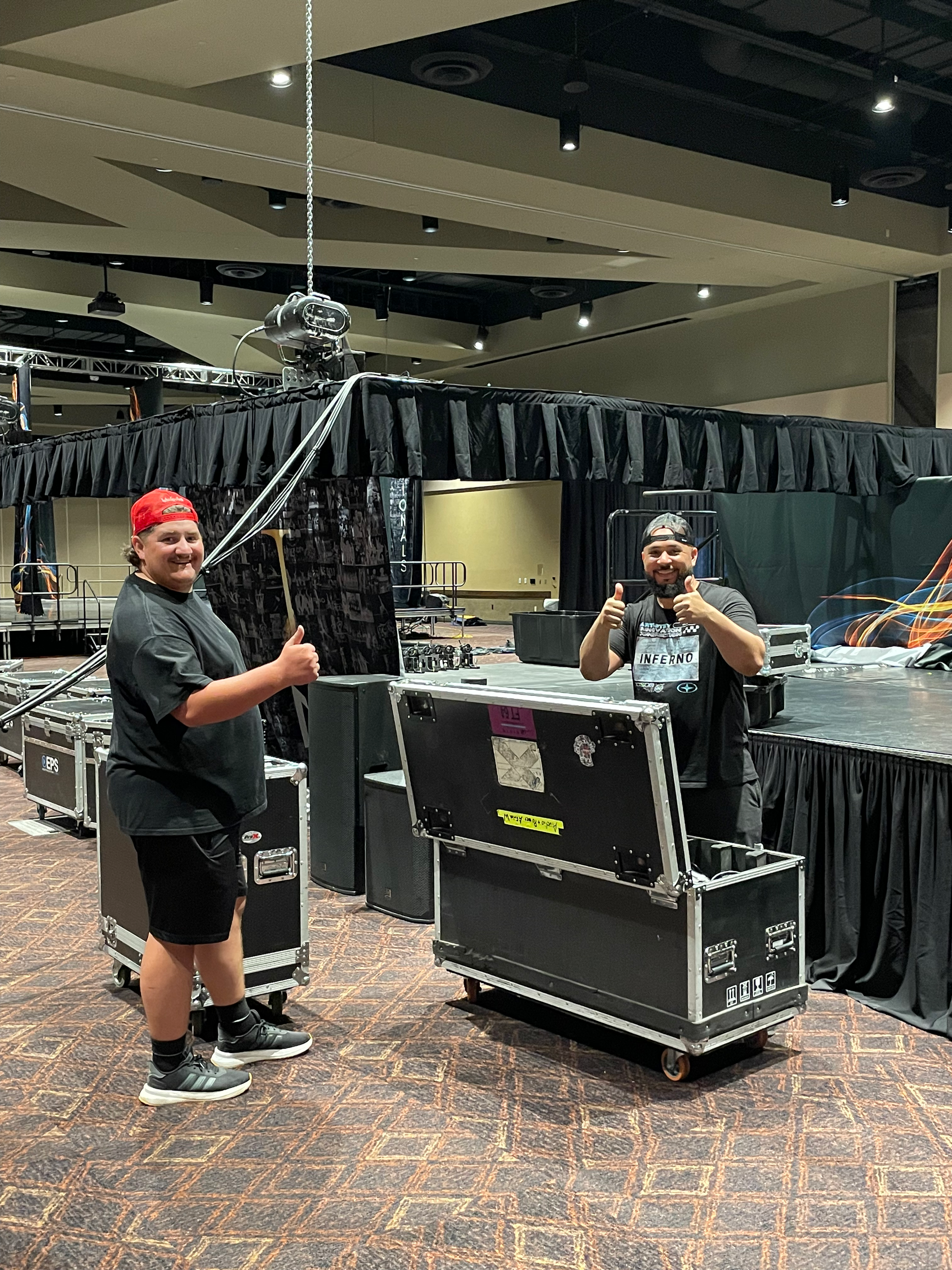 Two men standing in an indoor event space with a stage and black curtain backdrop, equipment cases, and technical gear. One man is wearing a red cap, black t-shirt, shorts, and sneakers, giving a thumbs up. The other man is wearing a black cap, black