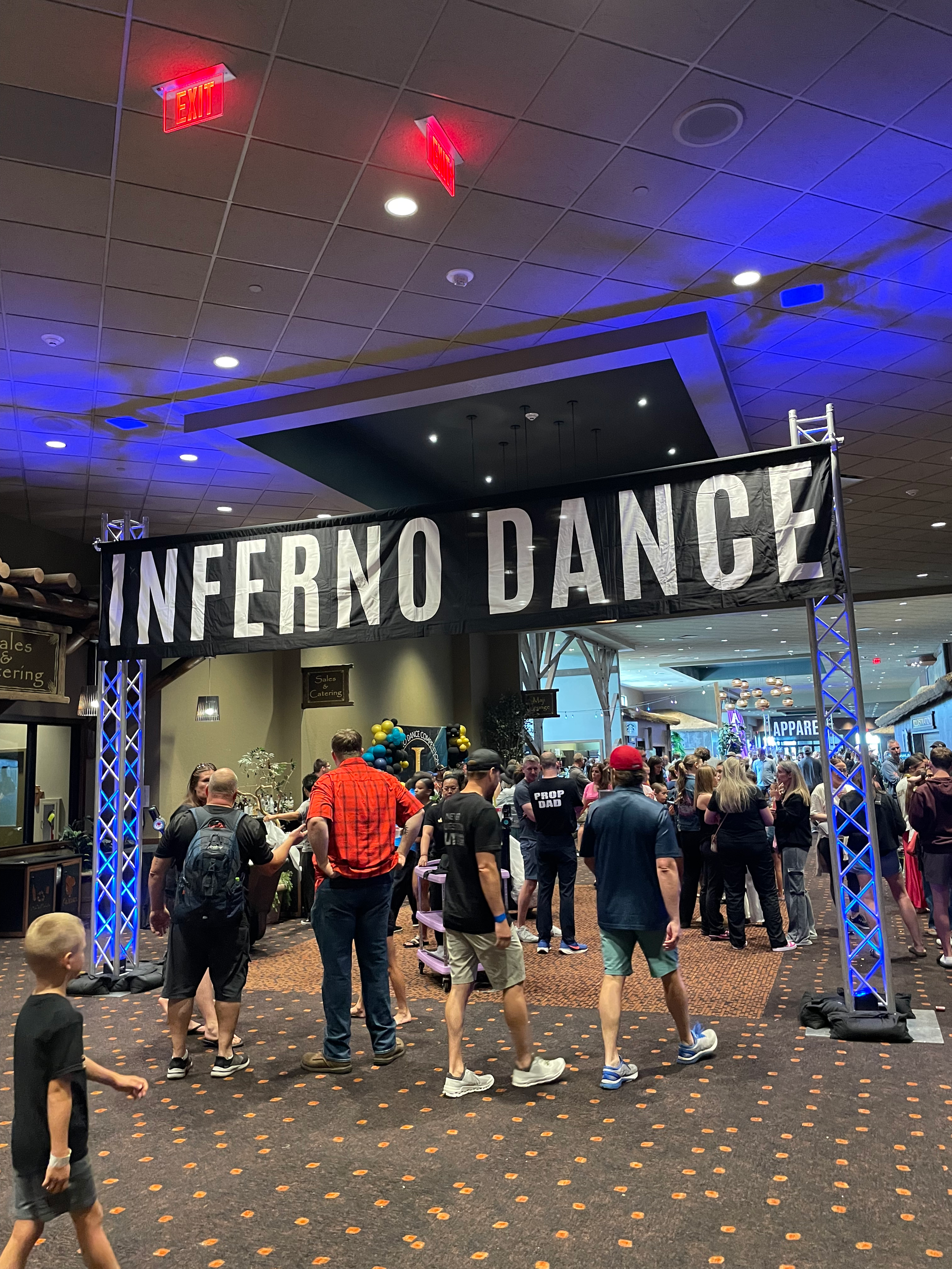 People gathered at an event with a large black banner reading 'INFERNO DANCE,' some people talking and walking around.