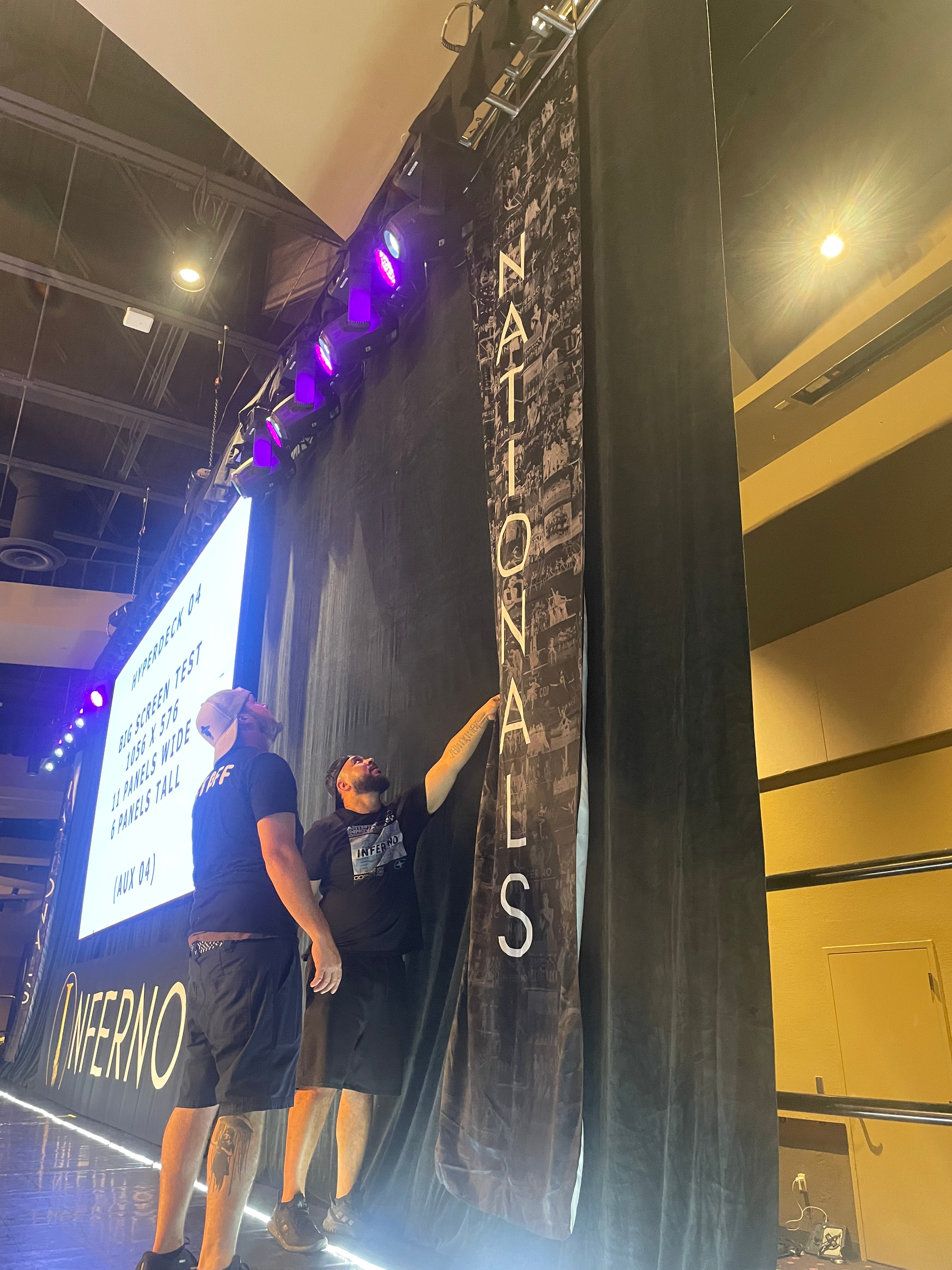 Two men at a tech event, one pointing at a large black banner that reads 'NADER,' while the other looks on. Behind them, a screen displays information about a big-screen test with panels.