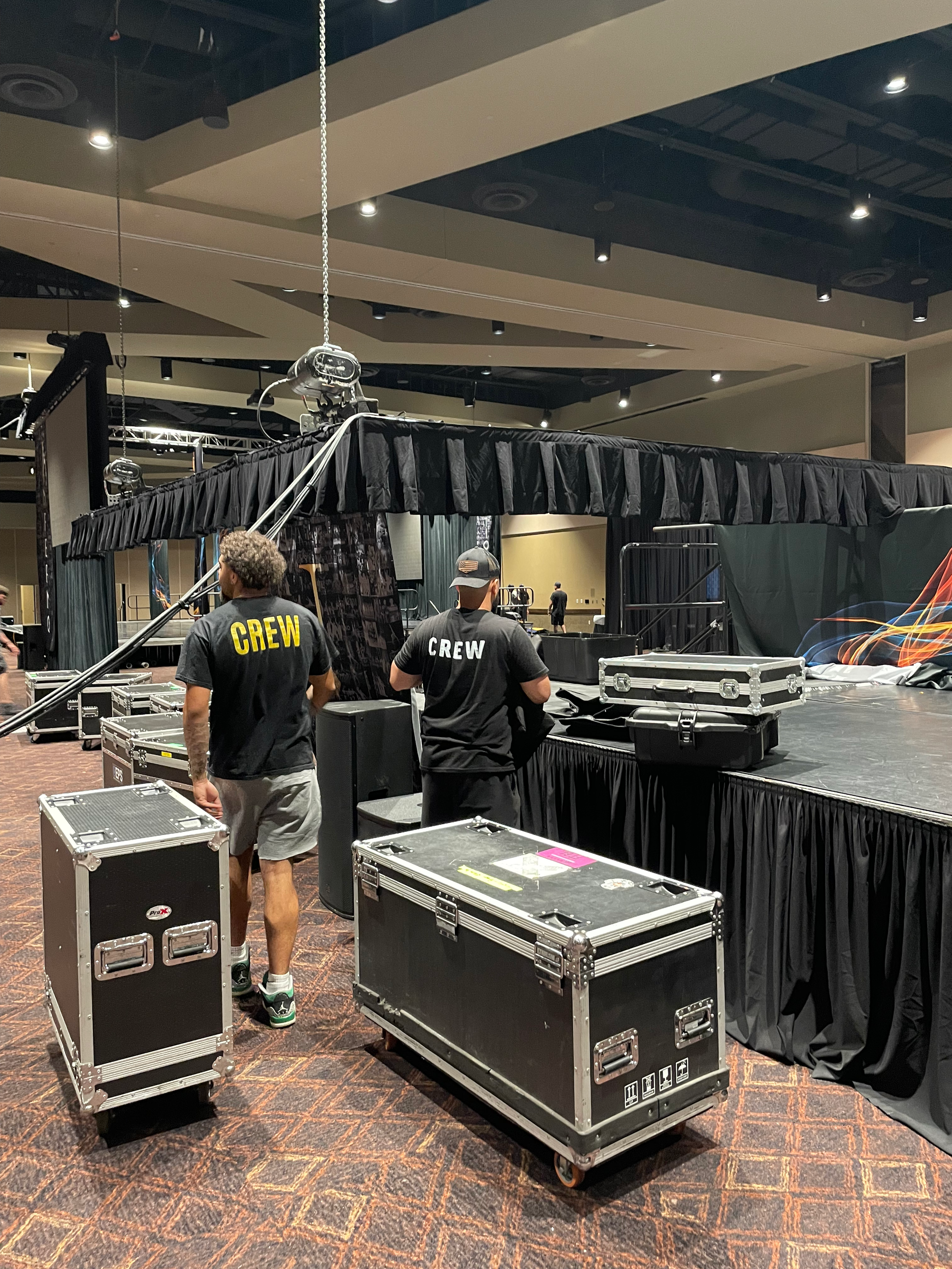 People setting up stage equipment in an indoor conference room, including crates with equipment, a stage with black curtains, and crew members working.
