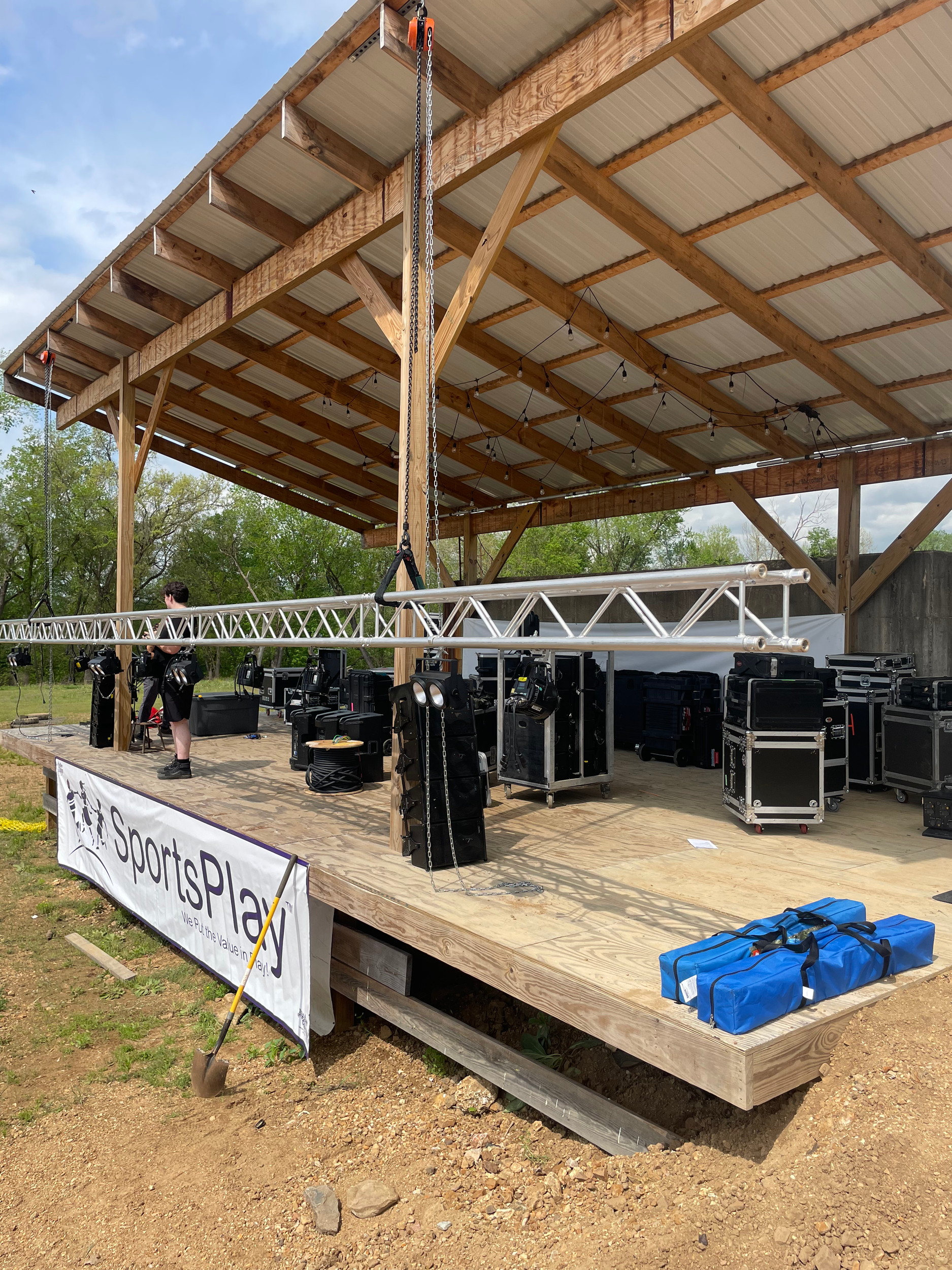 An outdoor stage under construction with a wooden structure, string lights, and various audio and lighting equipment. There is a person with a harness and a shovel on the stage, and a blue bag on the ground outside the stage.