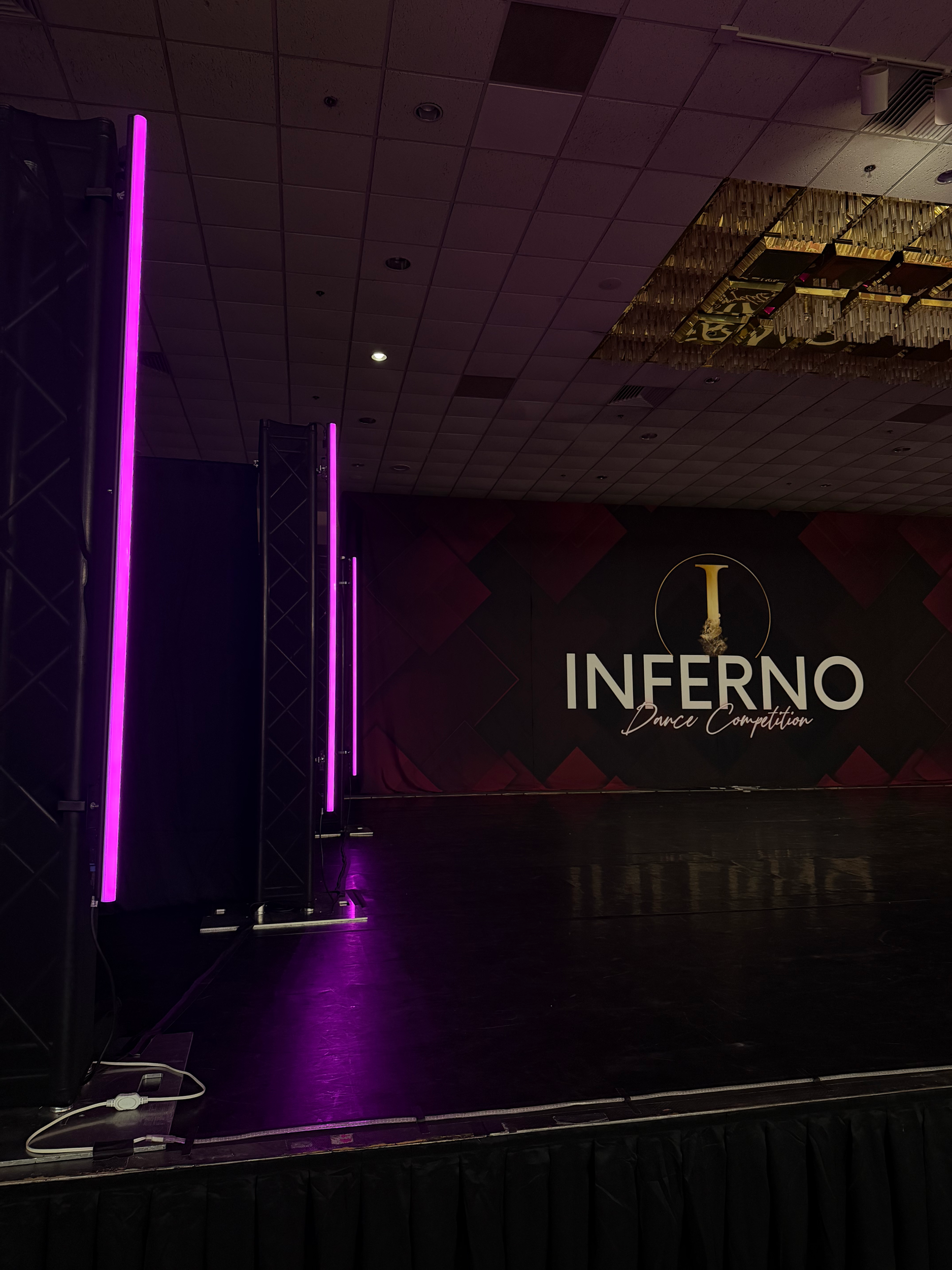 Stage setup for a dance competition called 'Inferno', featuring purple vertical lights, a dark stage with a shiny floor, and a decorative backdrop with a clock and the event name.