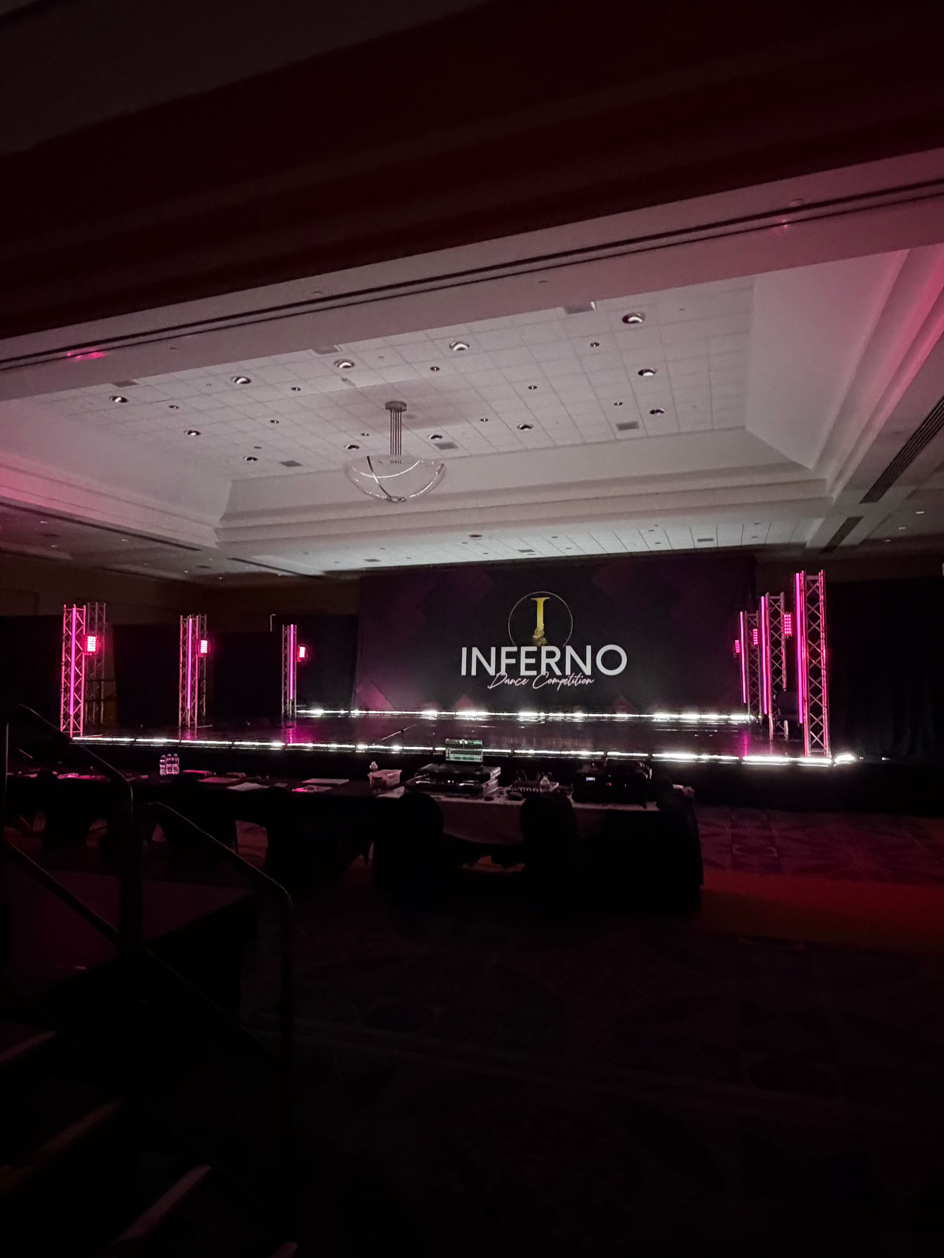 Empty event stage with LED lighting and a backdrop featuring the word "INFERNO" and the phrase "Dance Competition." The stage is lit with pink lights, with chairs and tables in the foreground.