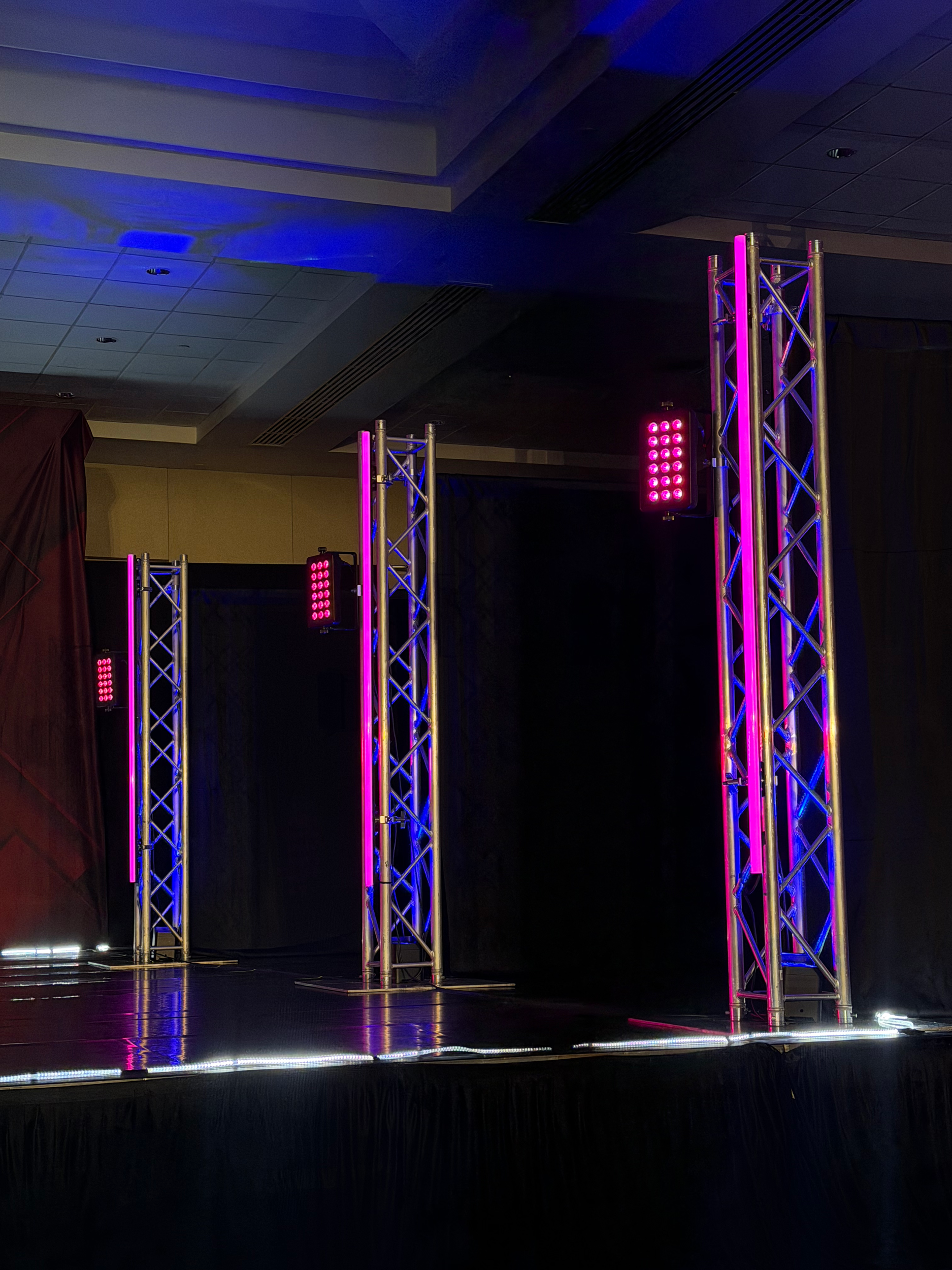 Stage setup with metal trusses illuminated with pink and purple lights, black curtains, and a dark ceiling with blue lighting reflections.