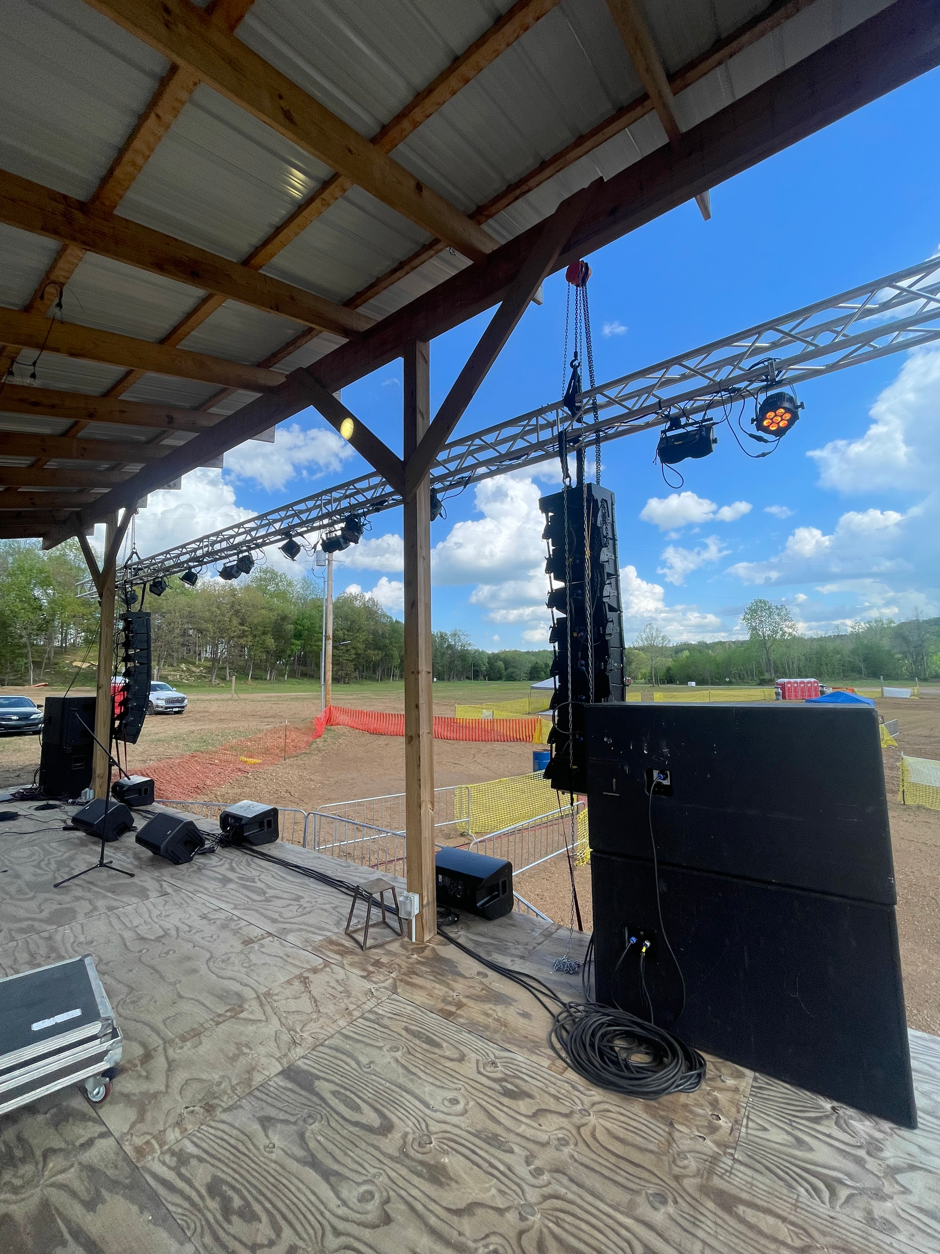 Outdoor stage setup with sound equipment, speakers, and lighting under a metal roof, overlooking a field with trees and blue sky.