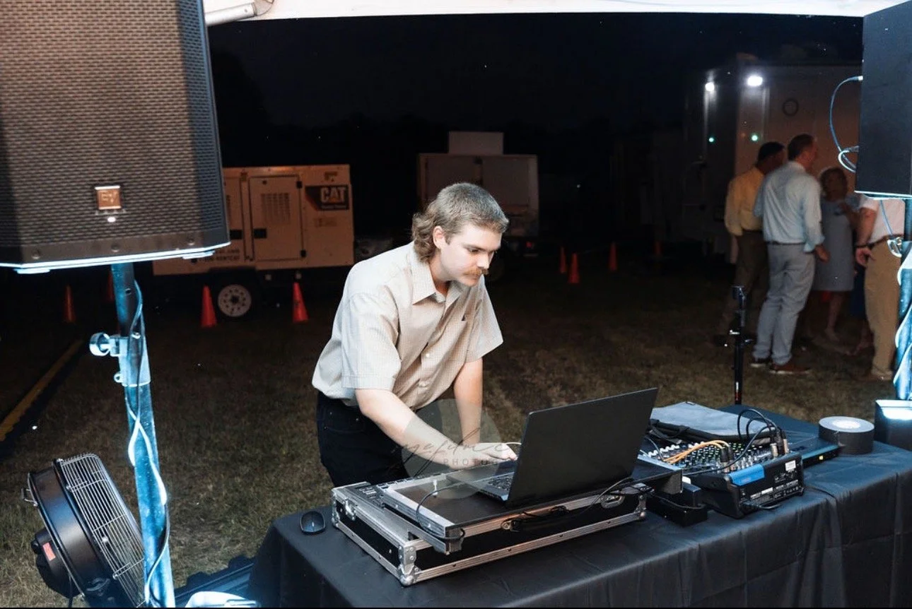 A DJ or music event technician at an outdoor at night, working on electronic music equipment including a laptop, mixer, and speakers, with people gathered in the background.