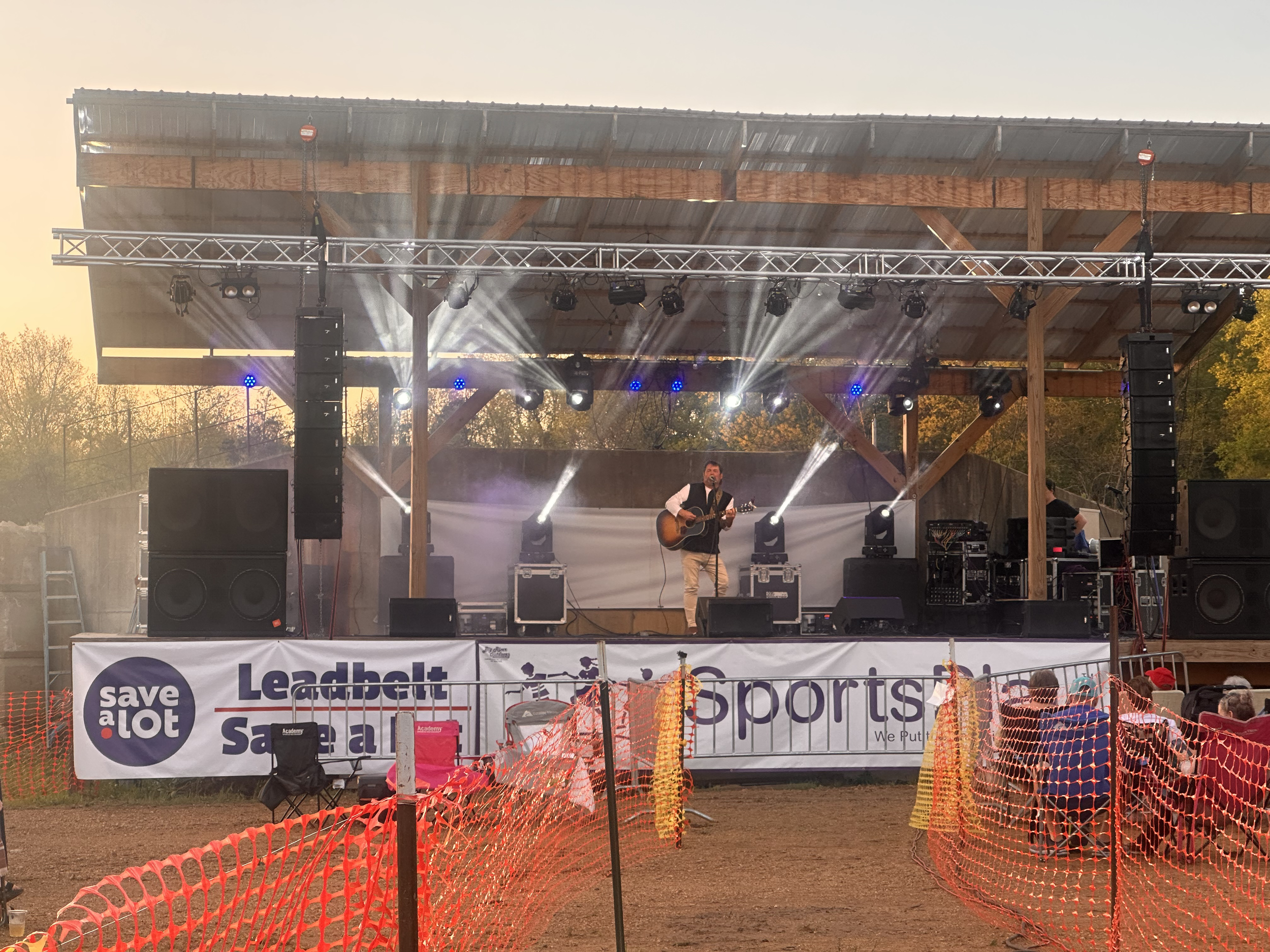 Stage with musician playing guitar under a large wooden canopy at an outdoor event during sunset. There are multiple music and lighting equipment and orange safety fencing in the foreground.