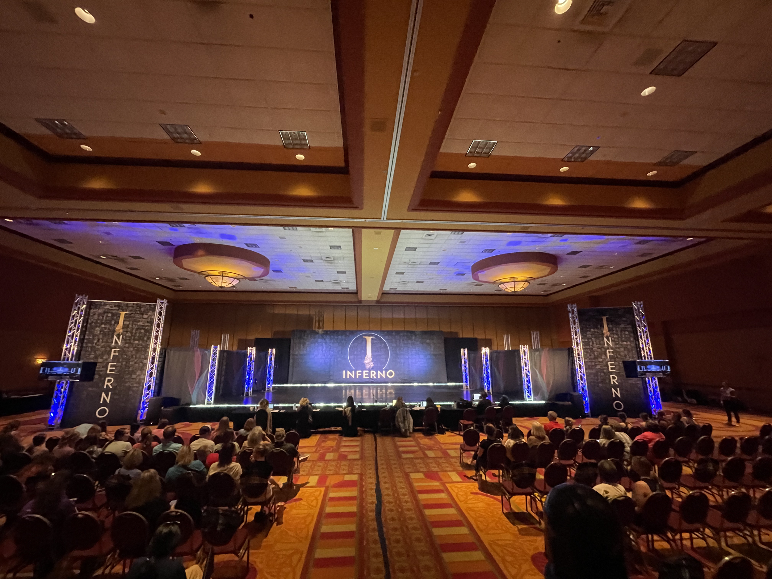 A large conference room with many attendees seated in chairs facing a stage with a black backdrop. The backdrop has gold text that reads 'Inferno' and some other smaller words, with vertical banners on either side spelling 'INFERNO'. The room has a h
