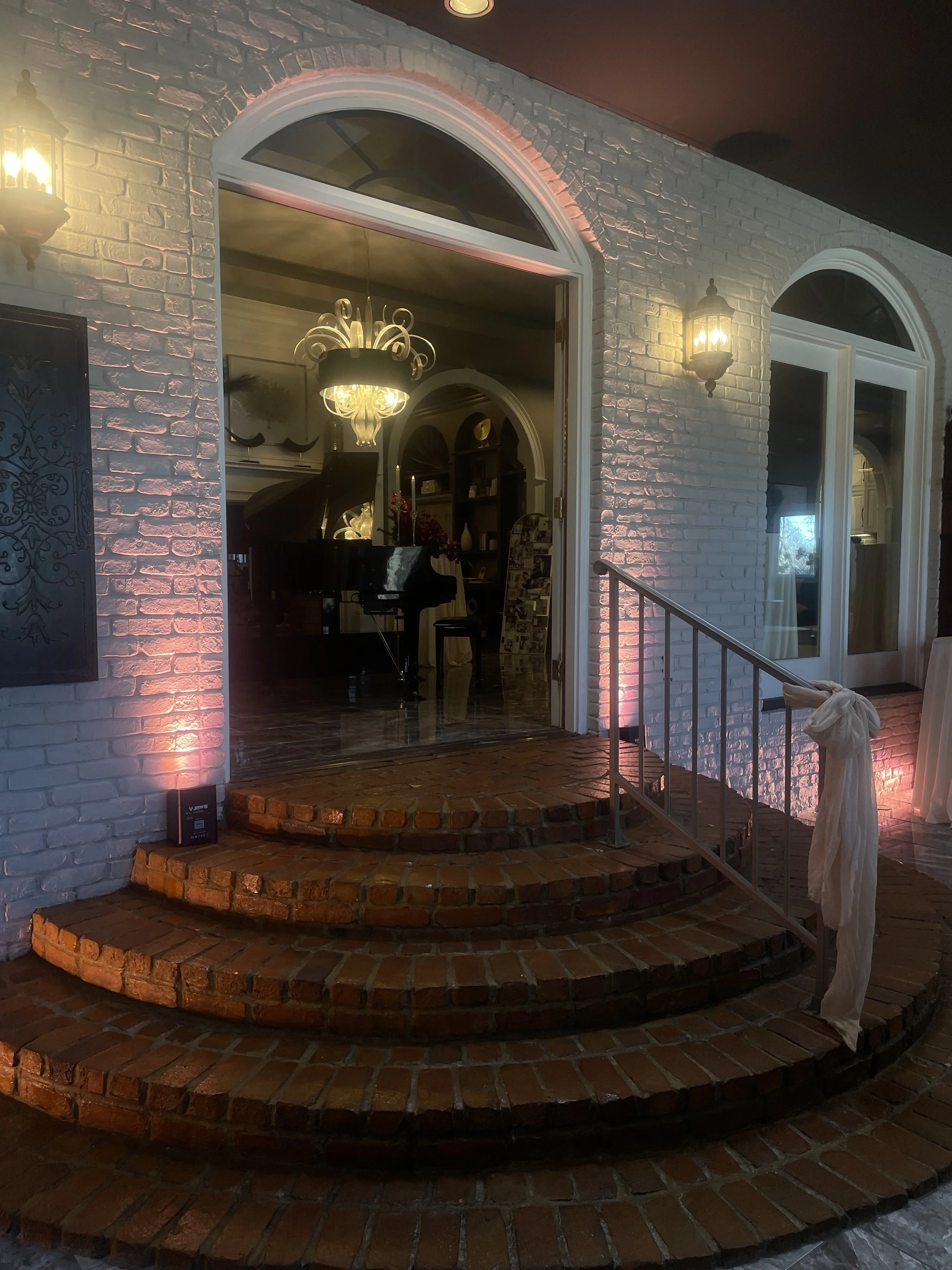 The image shows the exterior of a house with white brick walls and three arched windows. Two wall-mounted lantern lights illuminate the brick walls. A brick porch with two steps leads to an open doorway revealing a room inside with a black grand pian