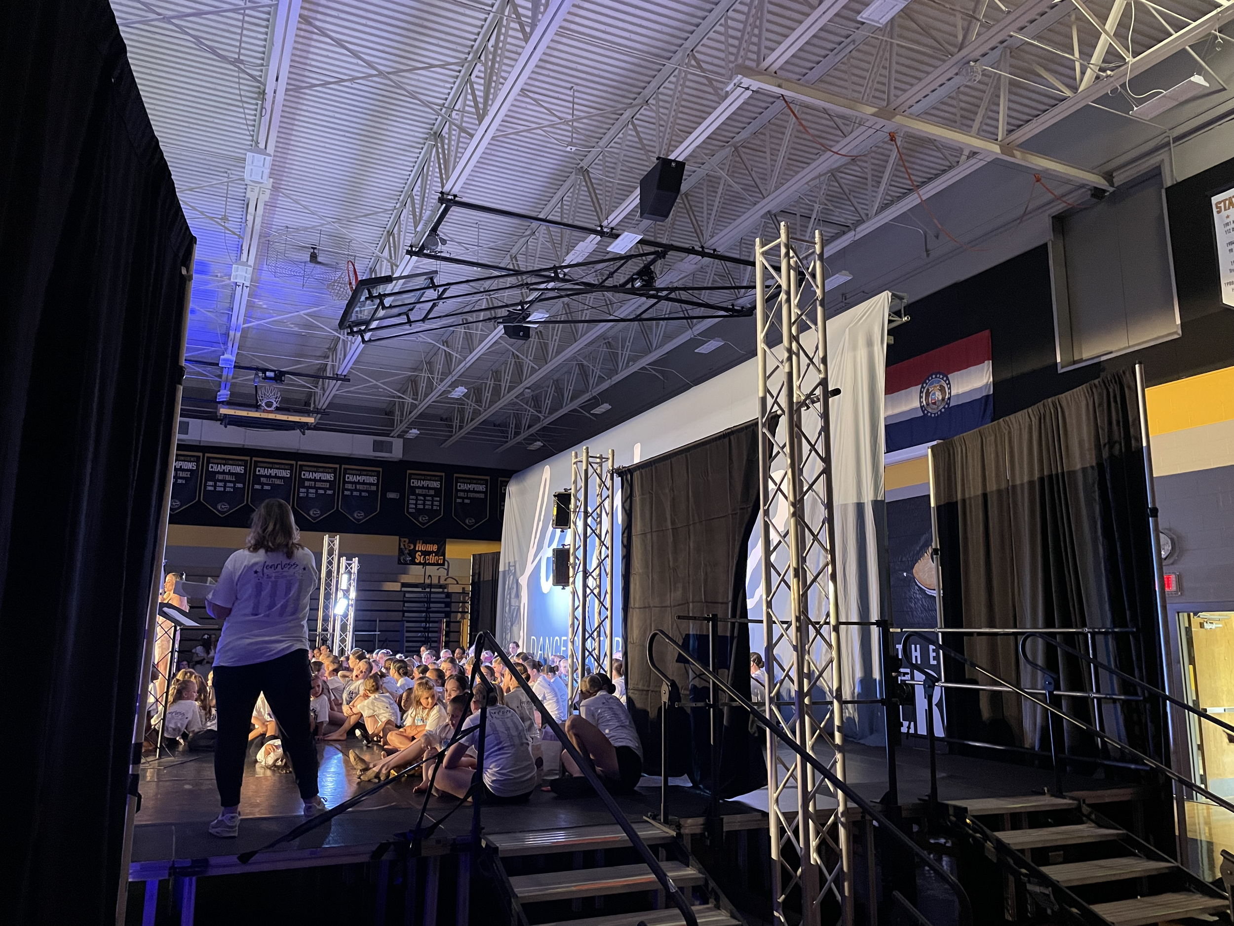 People sitting on the floor in a gymnasium with a crowd watching a performance or event, with banners and flags hanging on the wall and a stage setup.