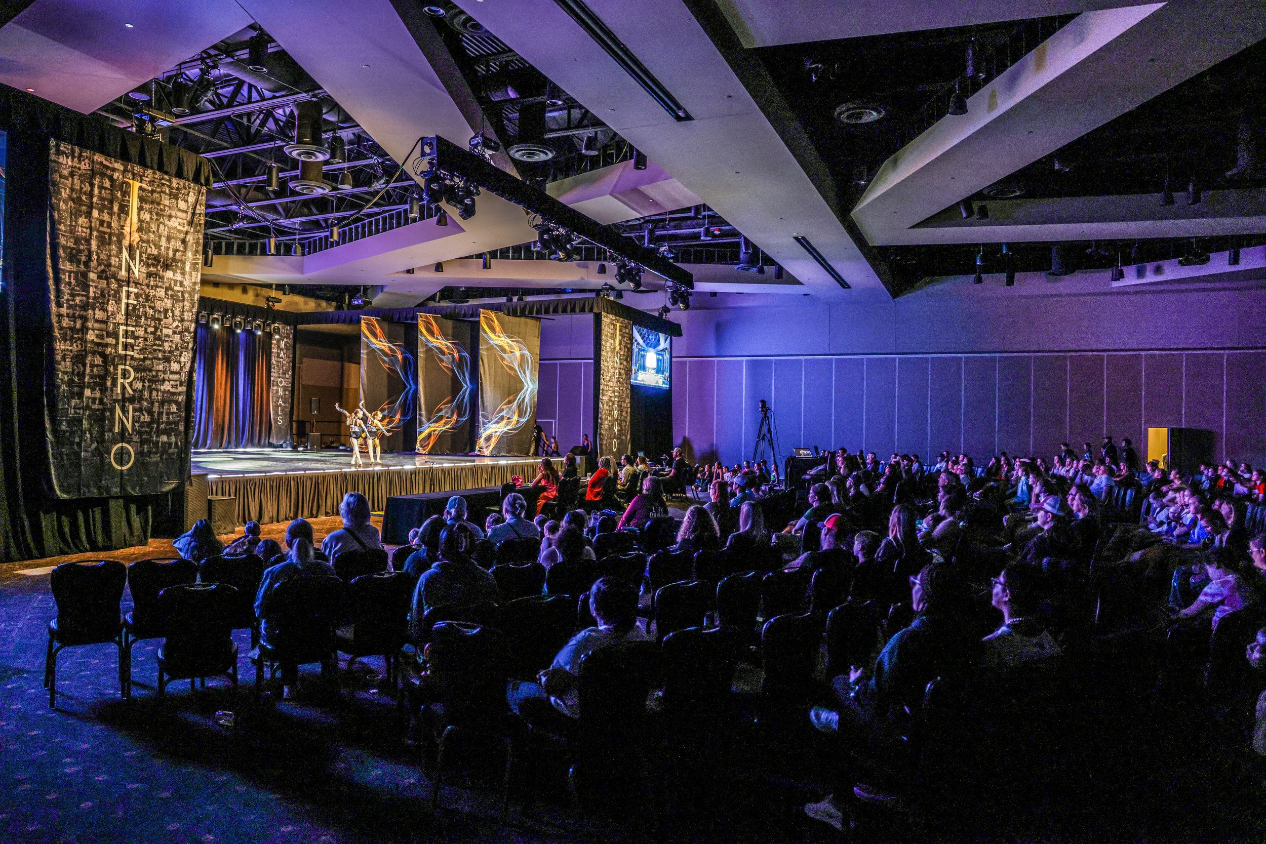 An indoor stage performance with dancers, audience seated, lighting effects, and large banners on the sides, in a darkened auditorium.