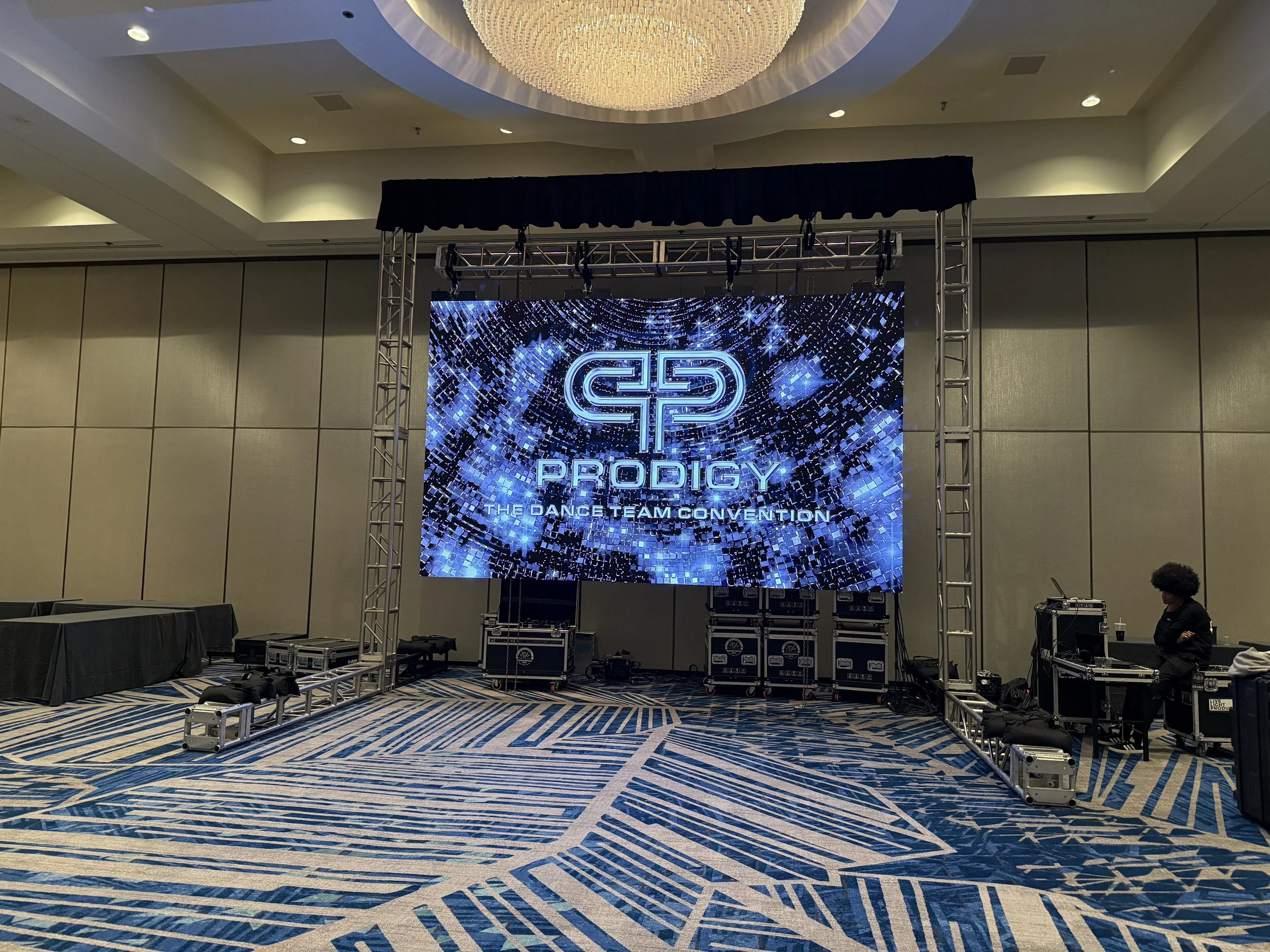 A large digital screen displays a logo with the word 'PRODIGY' and the phrase 'THE DANCE TEAM CONVENTION' beneath it, set up on a stage with professional lighting and sound equipment, in a spacious hotel conference room with patterned carpet and beig