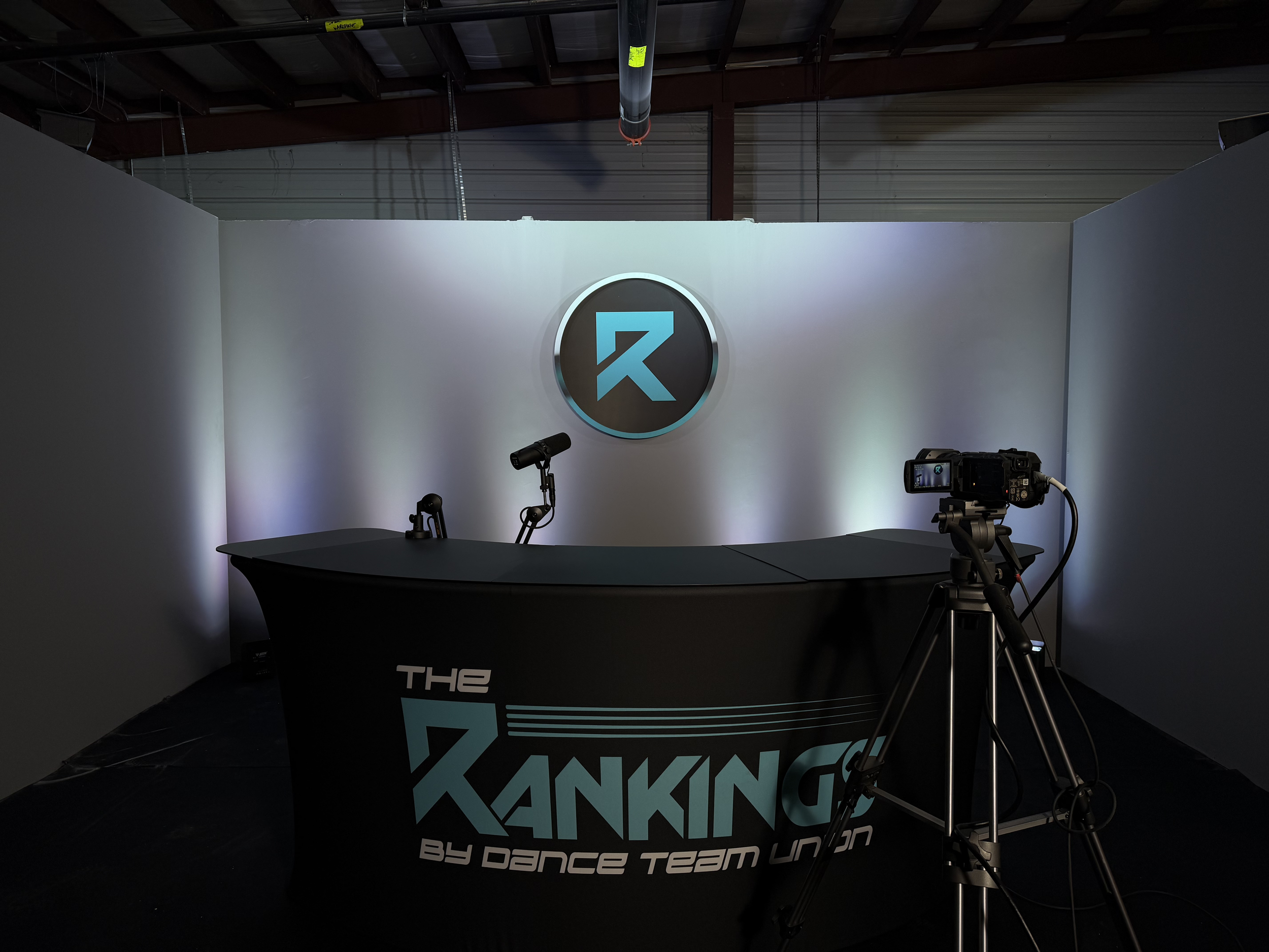 A photography setup with a black backdrop, spotlights, and a camera on a tripod, with a logo behind on a white wall reading "The Dancing by Dance Team Union" in blue and white.
