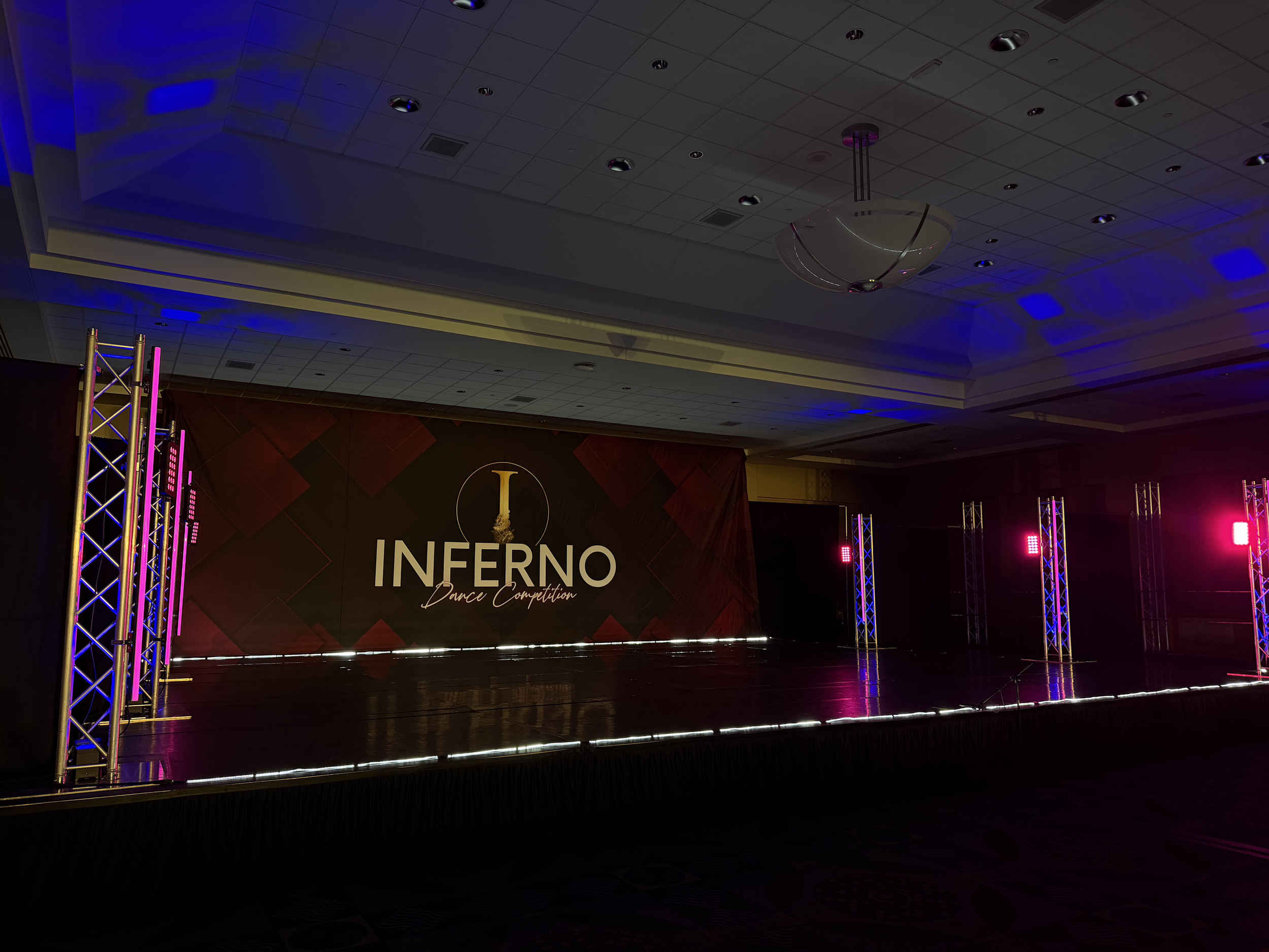 Stage with a banner displaying the words 'INFERNO Dance Competition', illuminated by purple and pink lighting, with truss structures on either side.