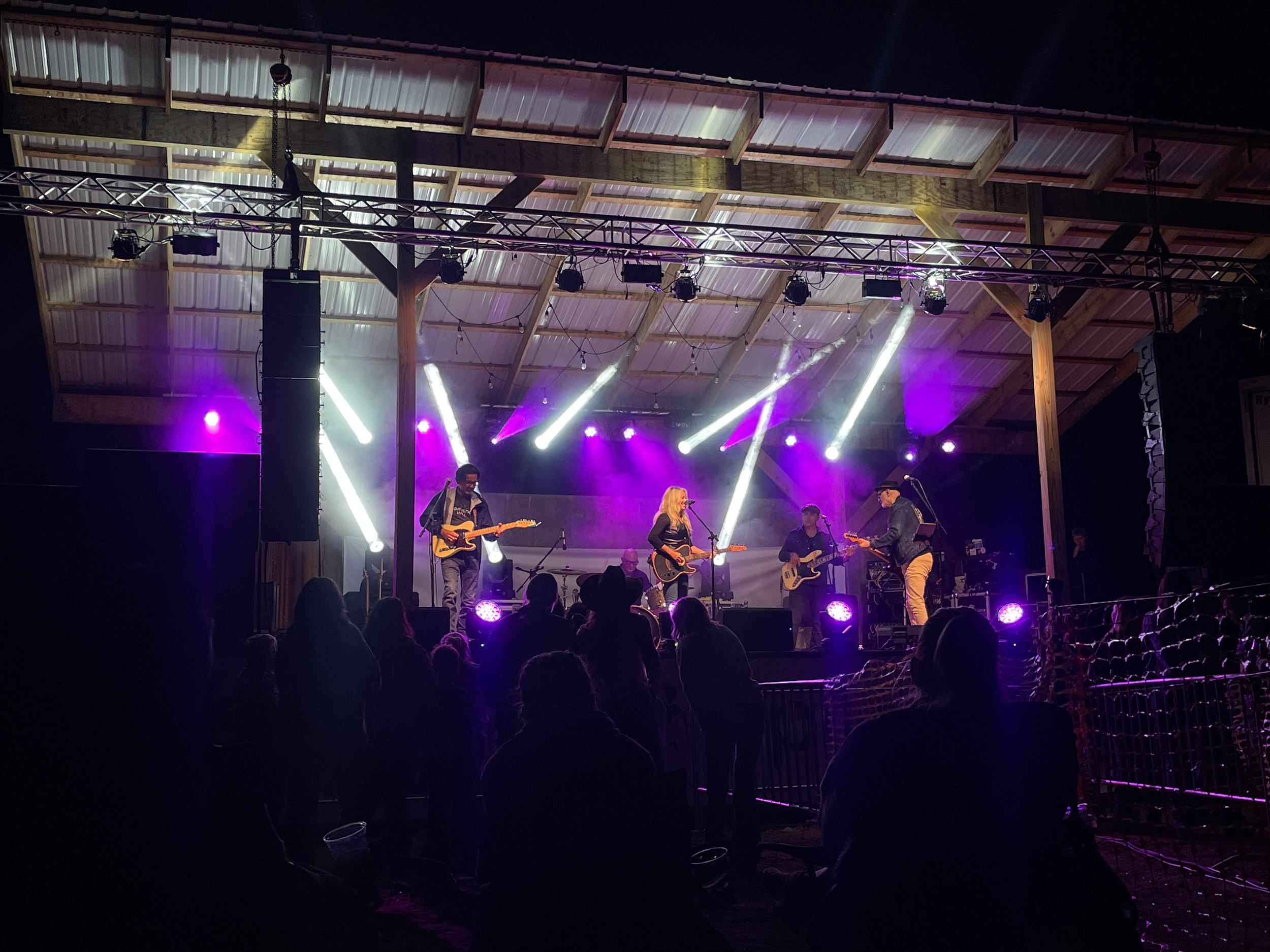 A live music concert with a band performing on stage under colorful stage lights, with an audience watching in a rustic outdoor venue at night.