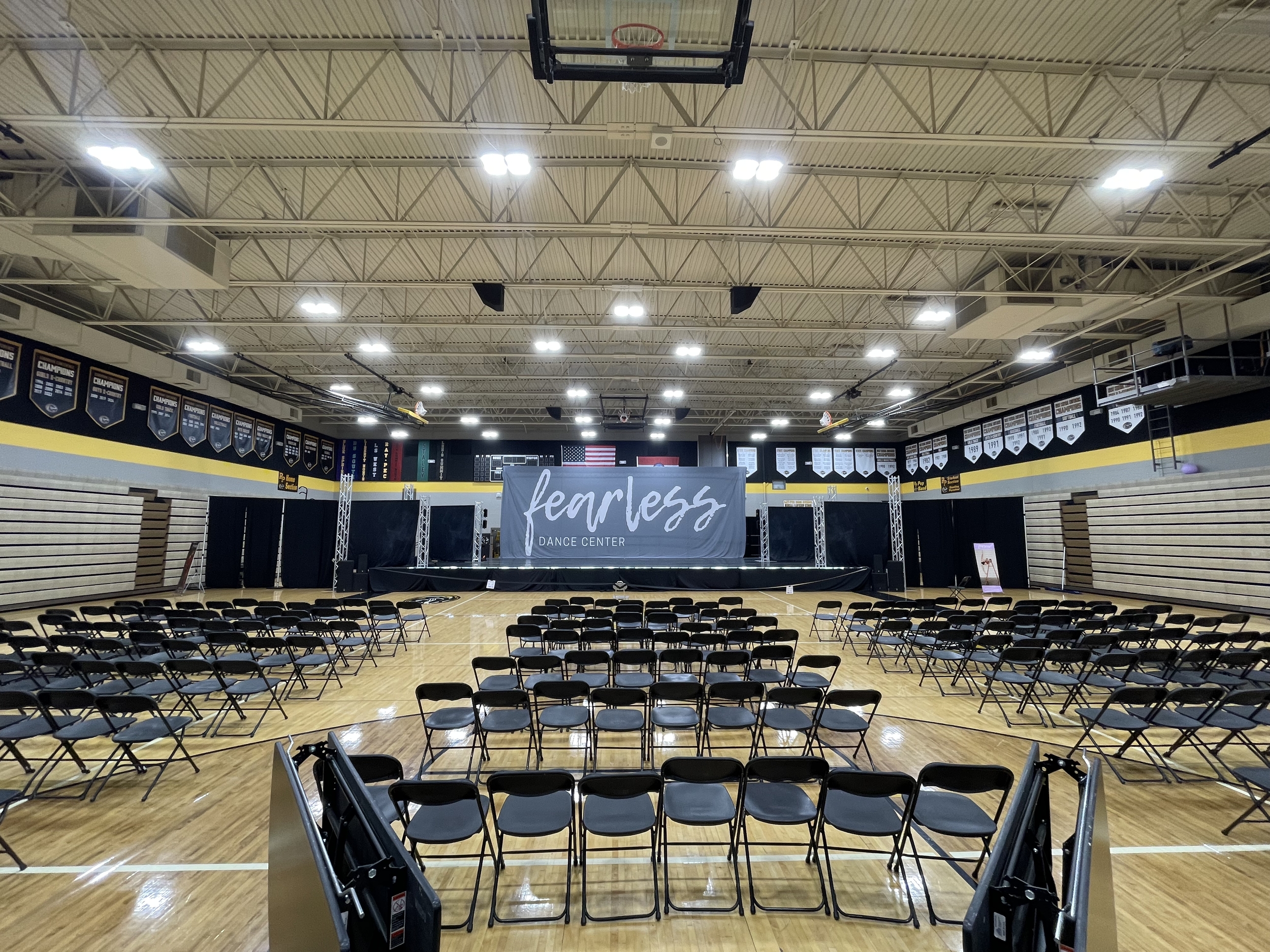 Interior of a gymnasium with a stage set up that has a sign reading 'fearless dance center', surrounded by rows of chairs for an audience, basketball hoops, banners hanging from the ceiling, and an American flag in the background.