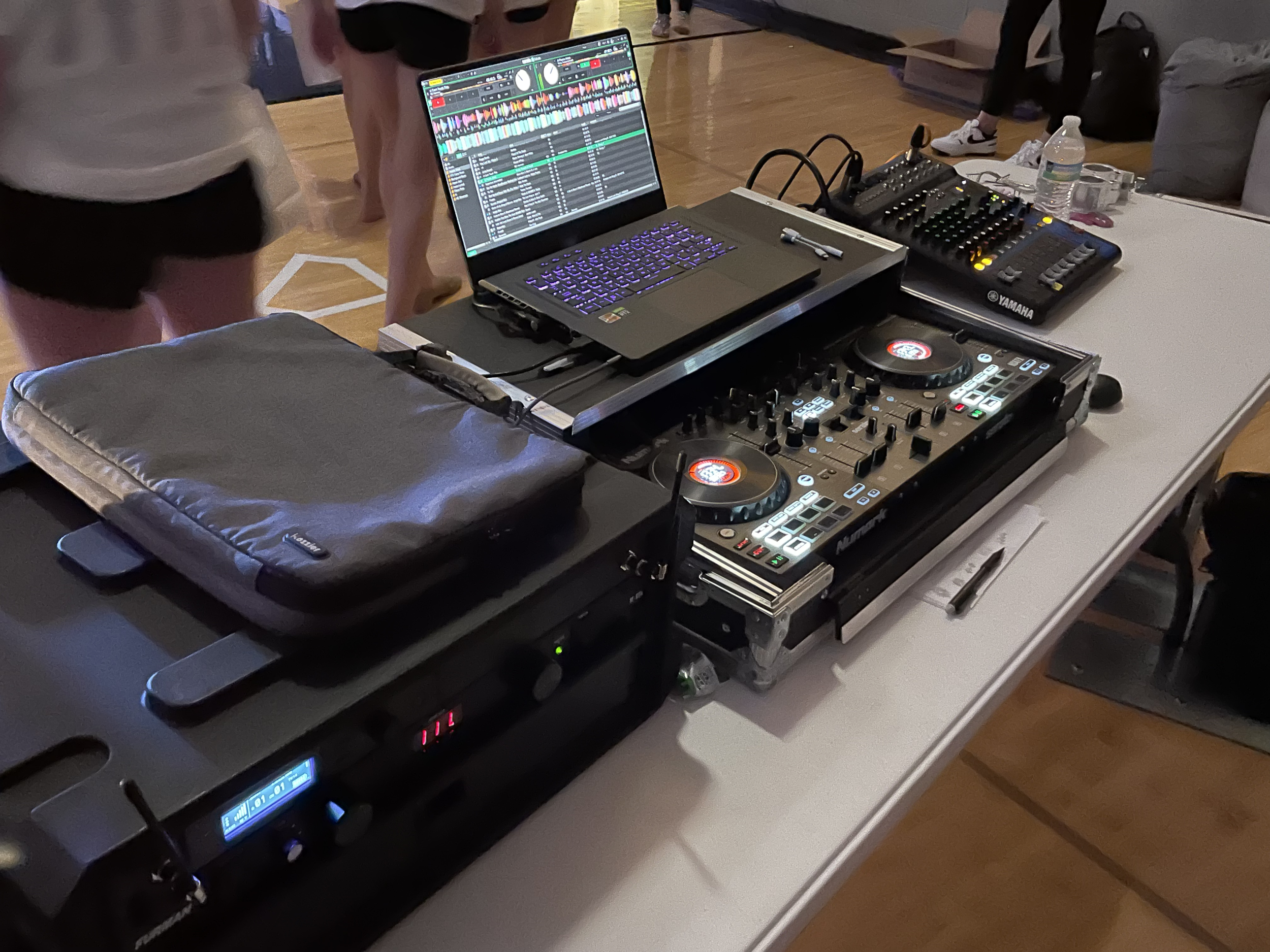 DJ equipment setup on a table includes a laptop with DJ software, a mixer, a controller, a water bottle, and some cables. People are standing in the background in a room with a wooden floor.