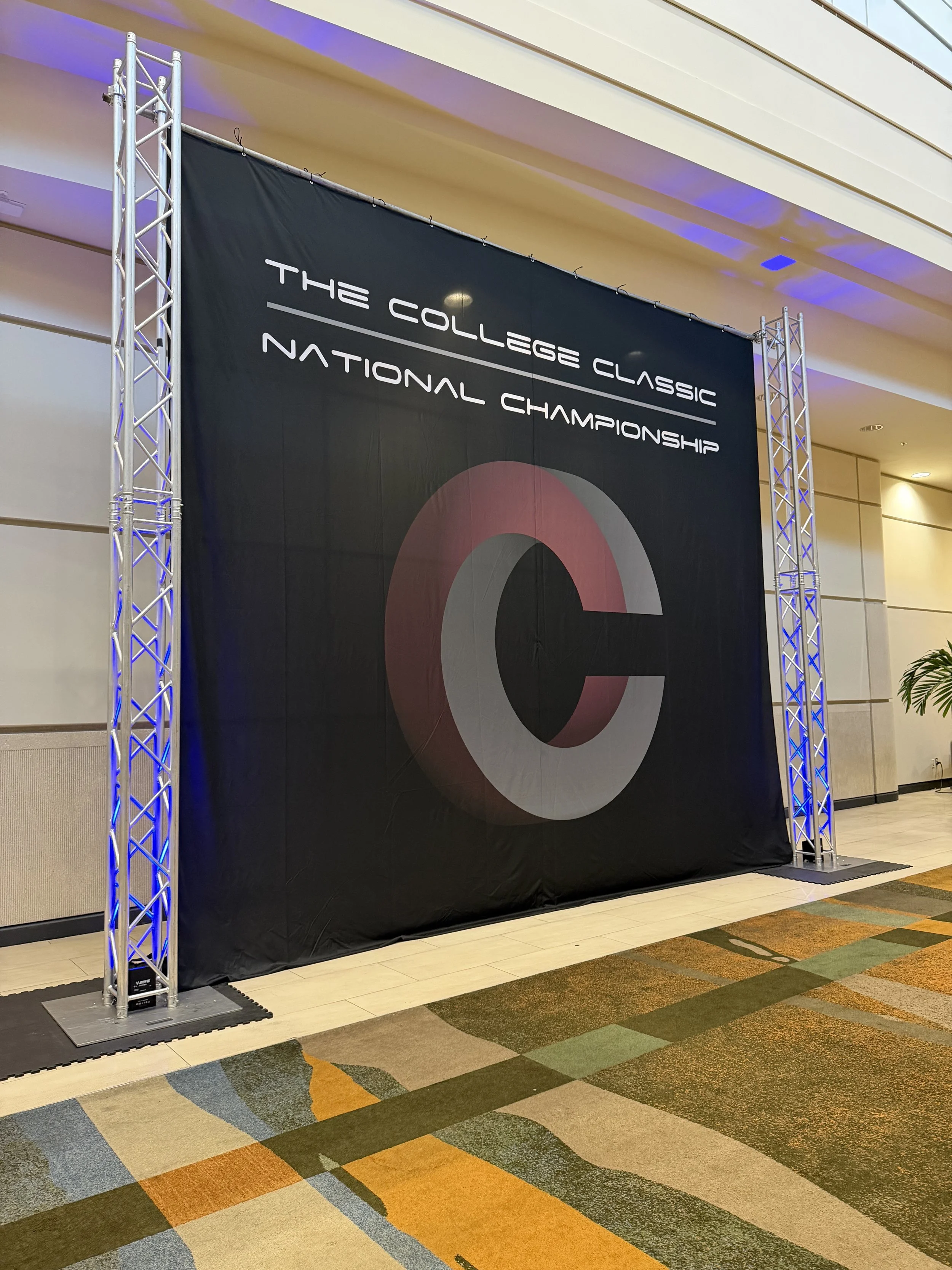 A large black banner with white text that reads 'The College Classic National Championship,' attached to a metal truss structure in an indoor venue with patterned carpeted floors and beige wall panels.
