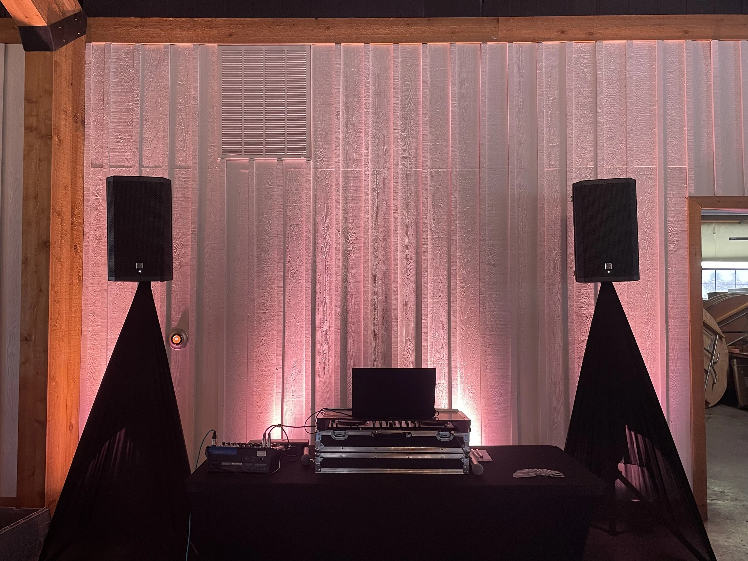 DJ equipment setup with two large black speakers on stands, a DJ mixer and turntable on a table, and colorful lighting on a wooden backdrop.