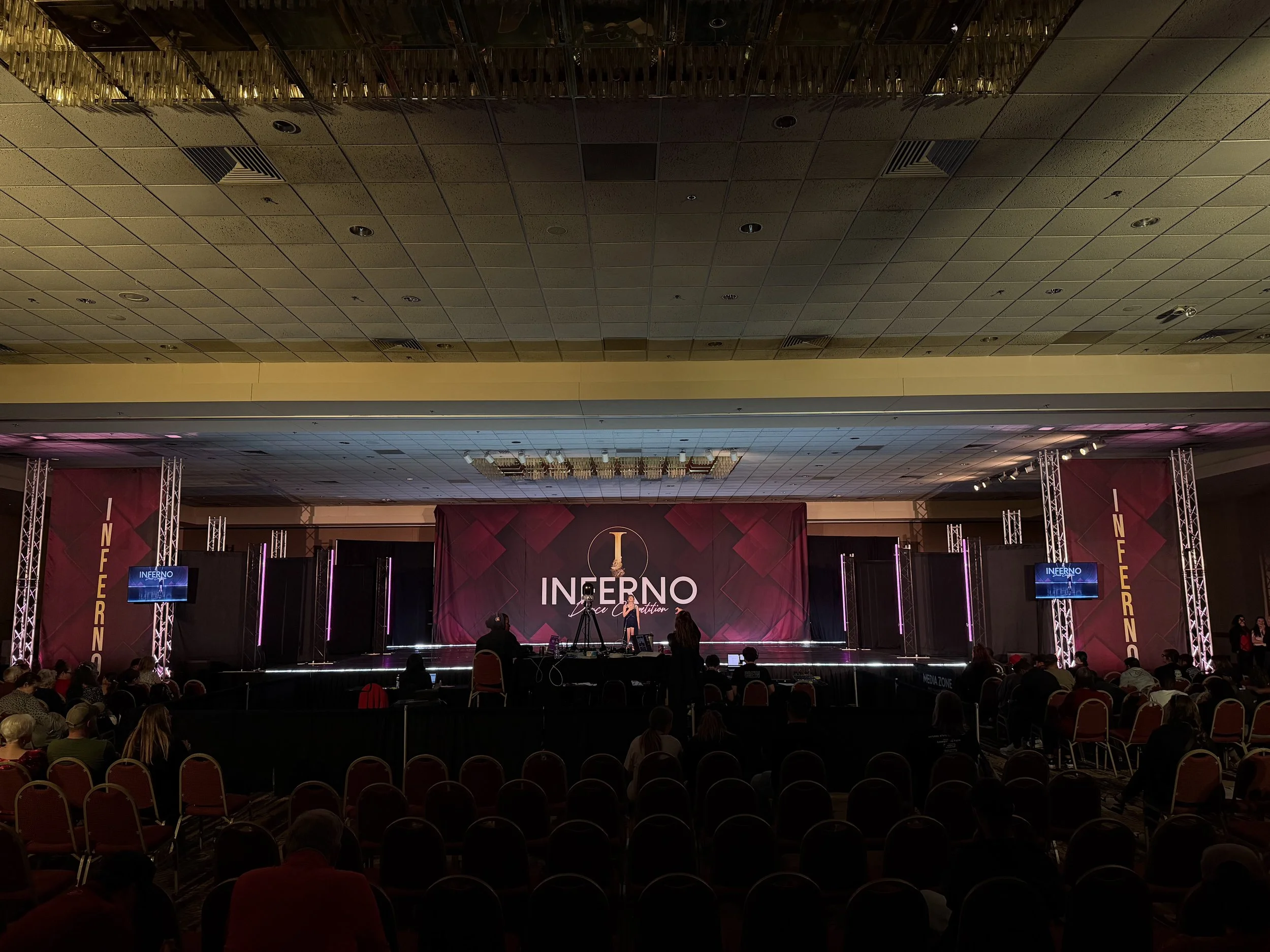 A stage set for a competition called Inferno, with a woman standing and speaking at a microphone. Audience members are seated in front of the stage. The stage features a large sign that says "INFERNO" and purple lighting.