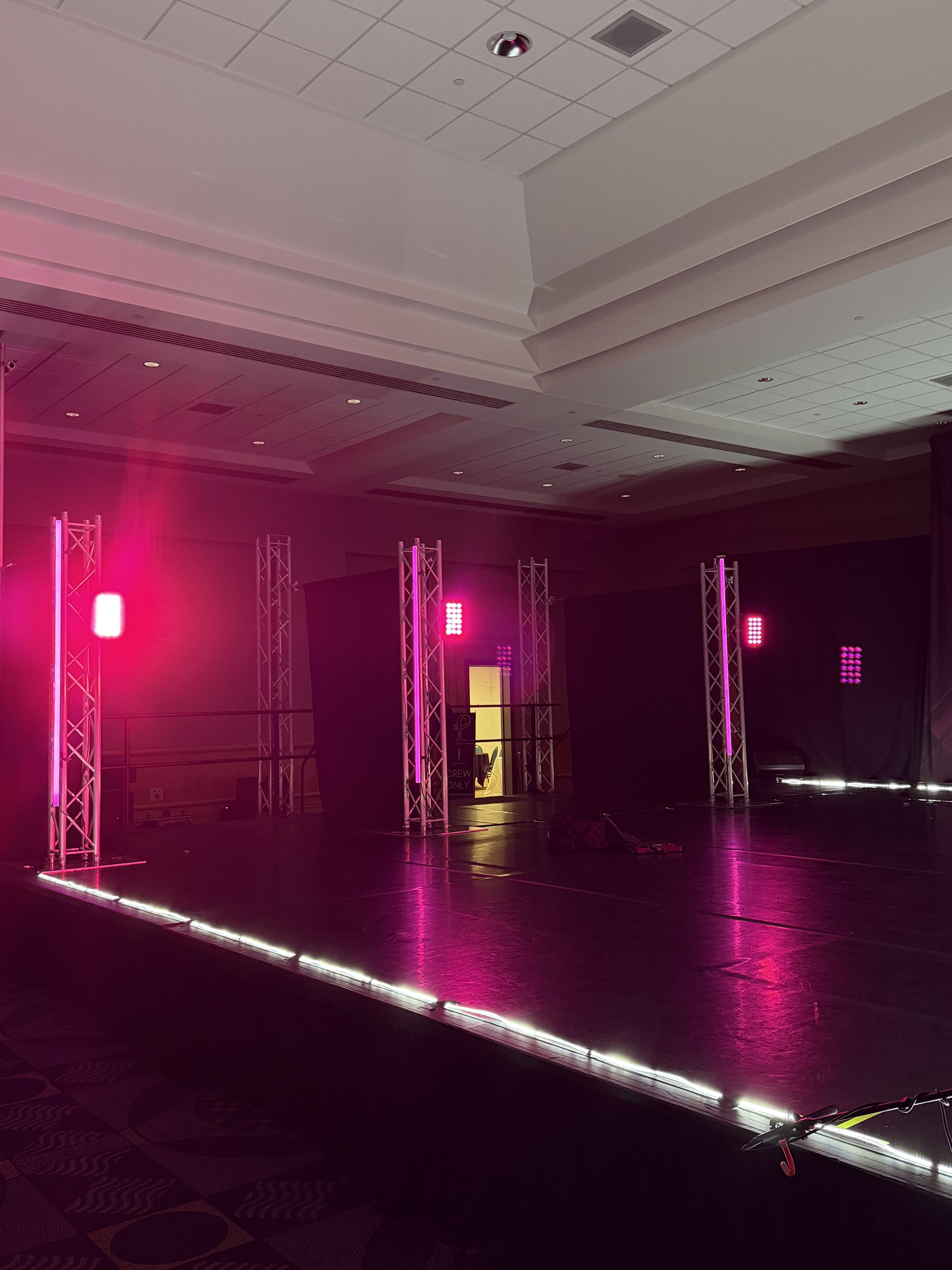 Empty event stage with pink lighting, metal trusses, and a dark backdrop in a large hall.