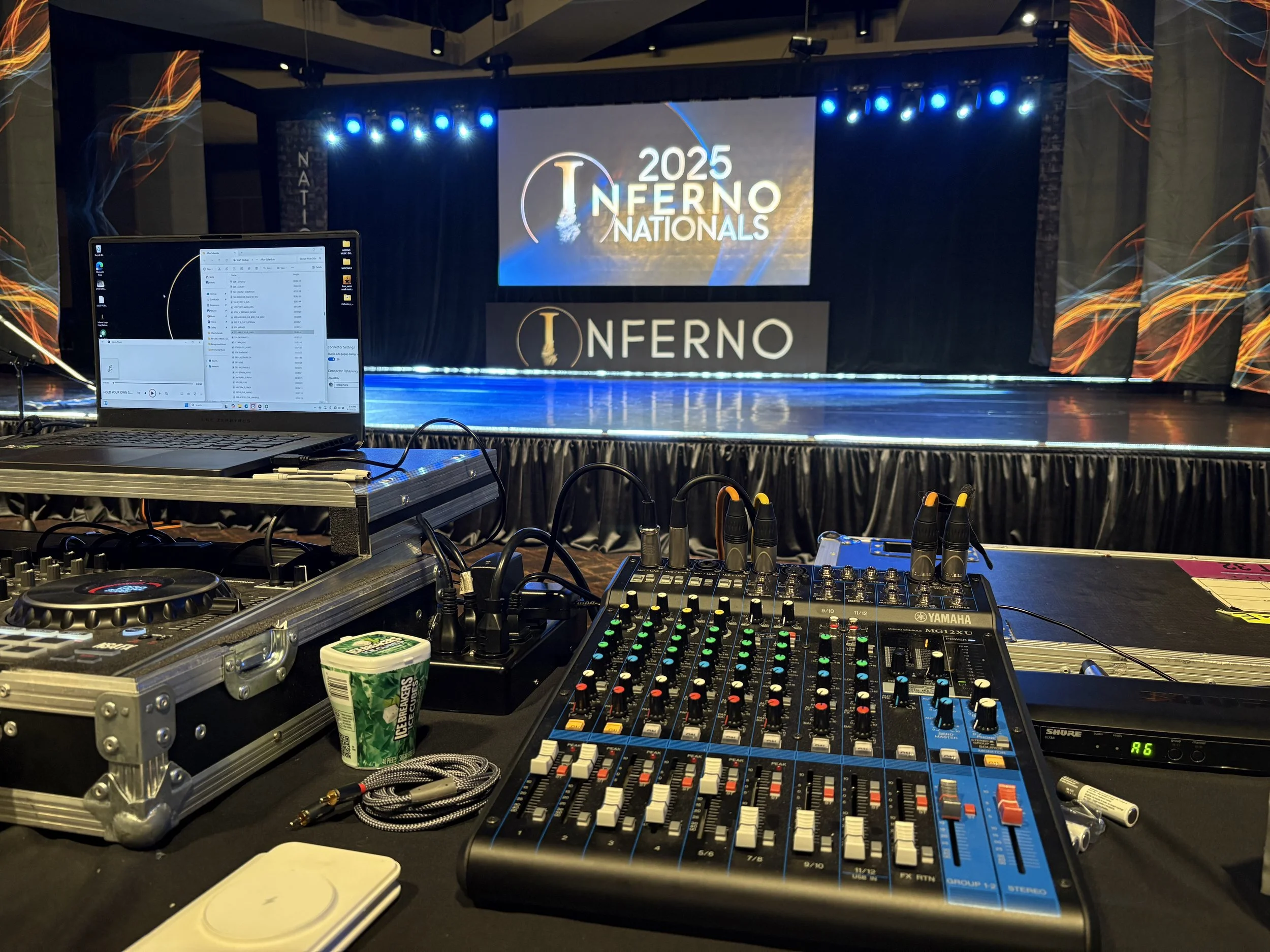 Sound mixing equipment on a table in front of a stage with a large screen displaying '2025 Inferno Nationals' in a darkened room.