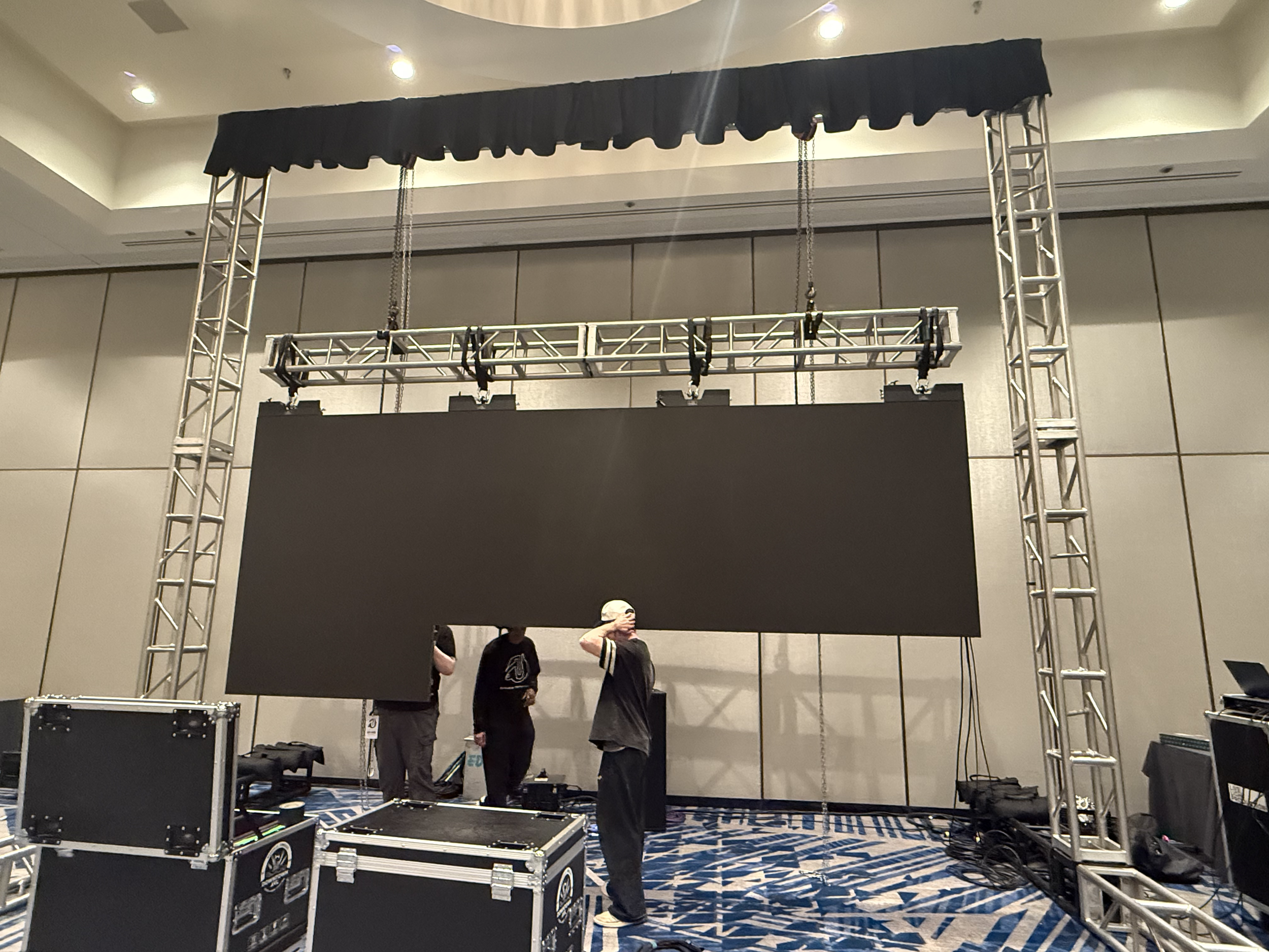 People setting up a stage with a black backdrop, metal truss structures, and hanging chains in a conference or event space.