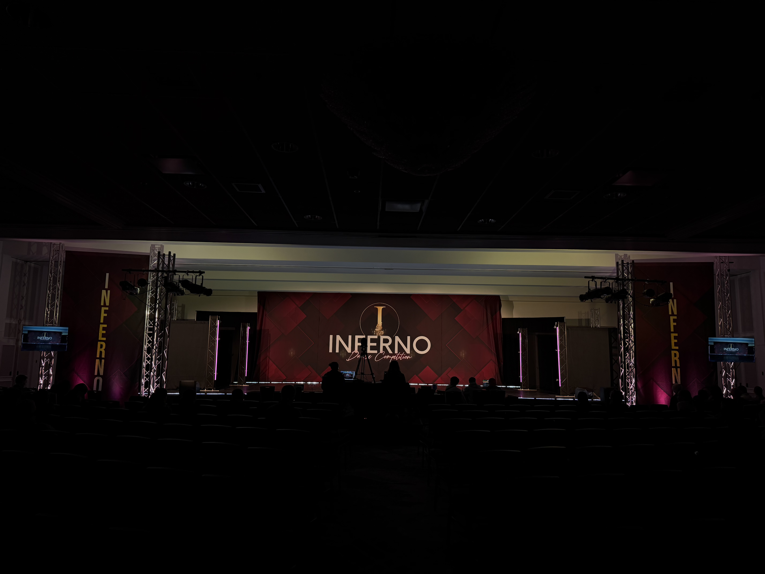 An indoor stage with a large screen displaying the word 'INFERNO' and a stylized image, with stage lighting and seating audience visible in shadows.