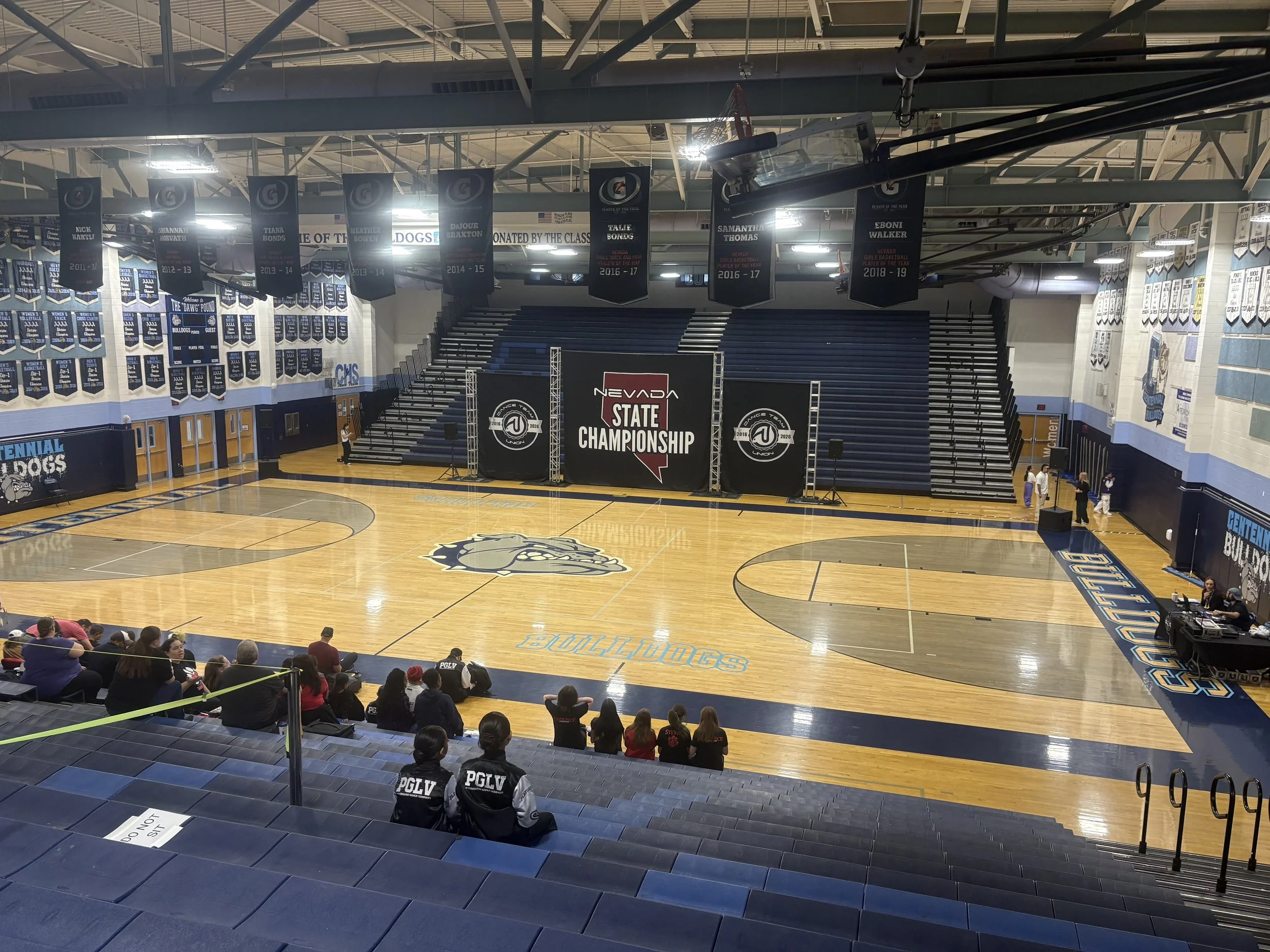 Inside a gymnasium with a basketball court, cheering fans sitting on seats, banners hanging from the ceiling, and a stage with a large Nevada State Championship sign. The gym has blue and white colors with banners on the walls.