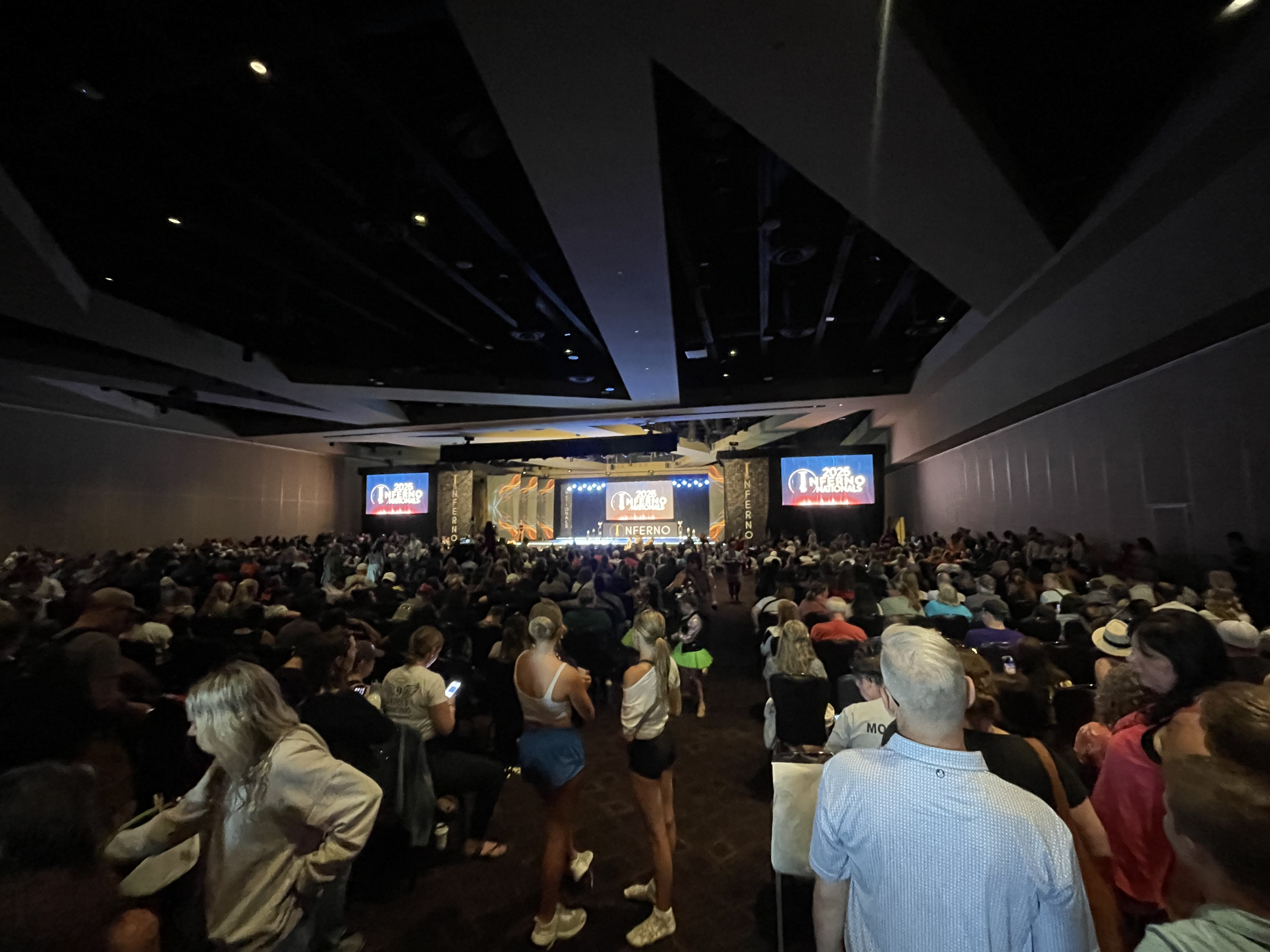 Large indoor conference or concert hall filled with people seated and facing a stage with multiple screens displaying the event's logo and text.