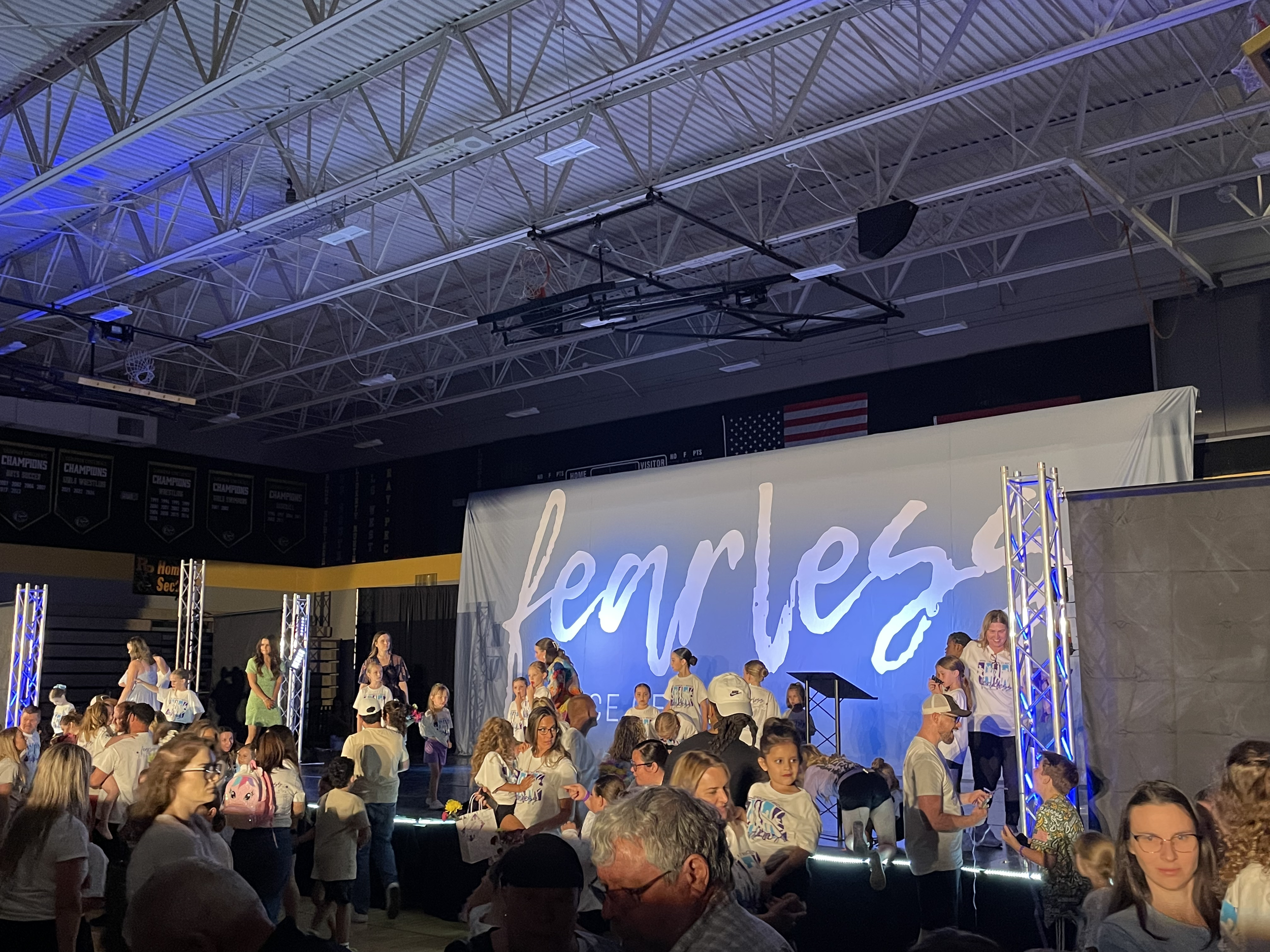 Indoor event with a large crowd of people, many children, gathered in front of a stage with a large banner that reads 'Remember'. The ceiling has visible metal beams, lighting, and audio equipment, with an American flag hanging on the wall.