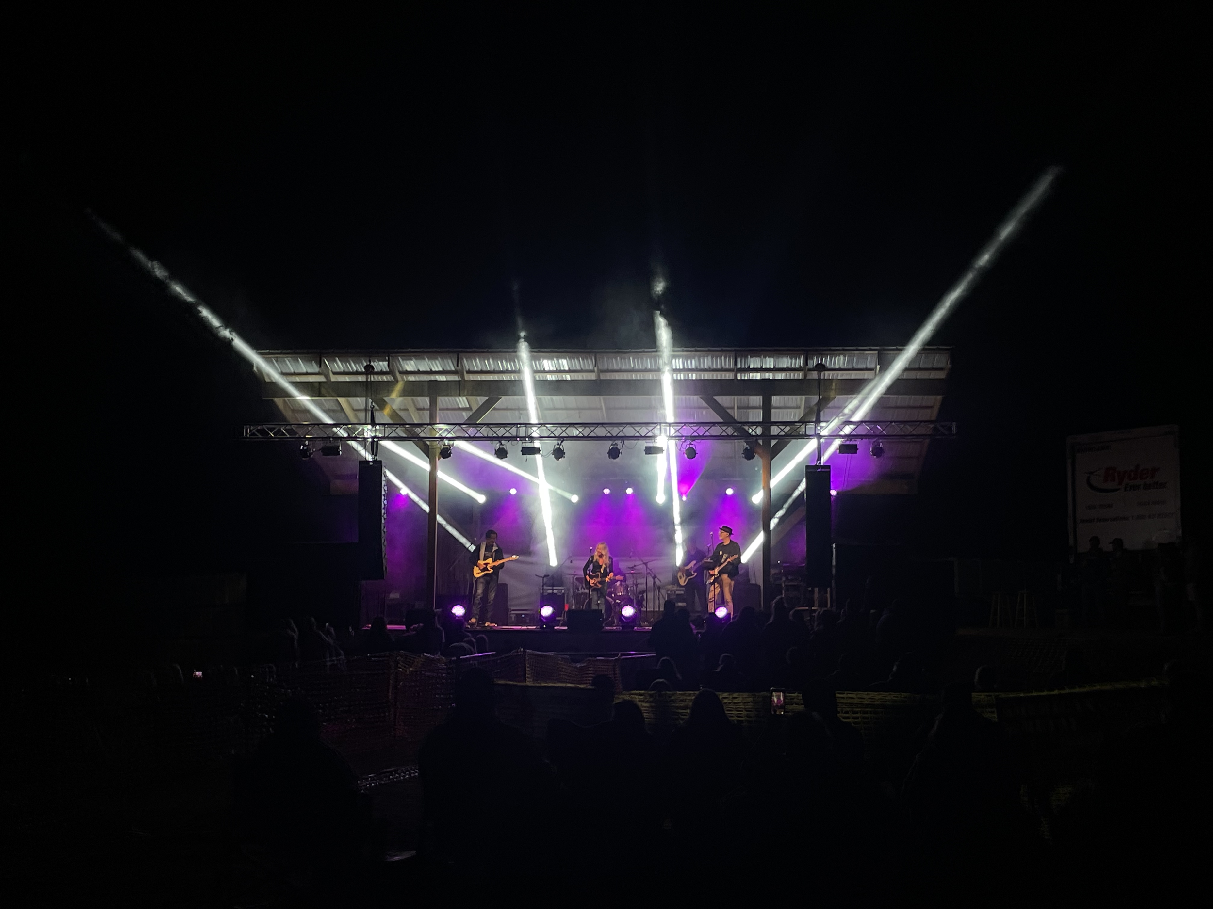Nighttime outdoor concert with a band playing on stage with purple and white lighting, audience silhouettes in foreground.