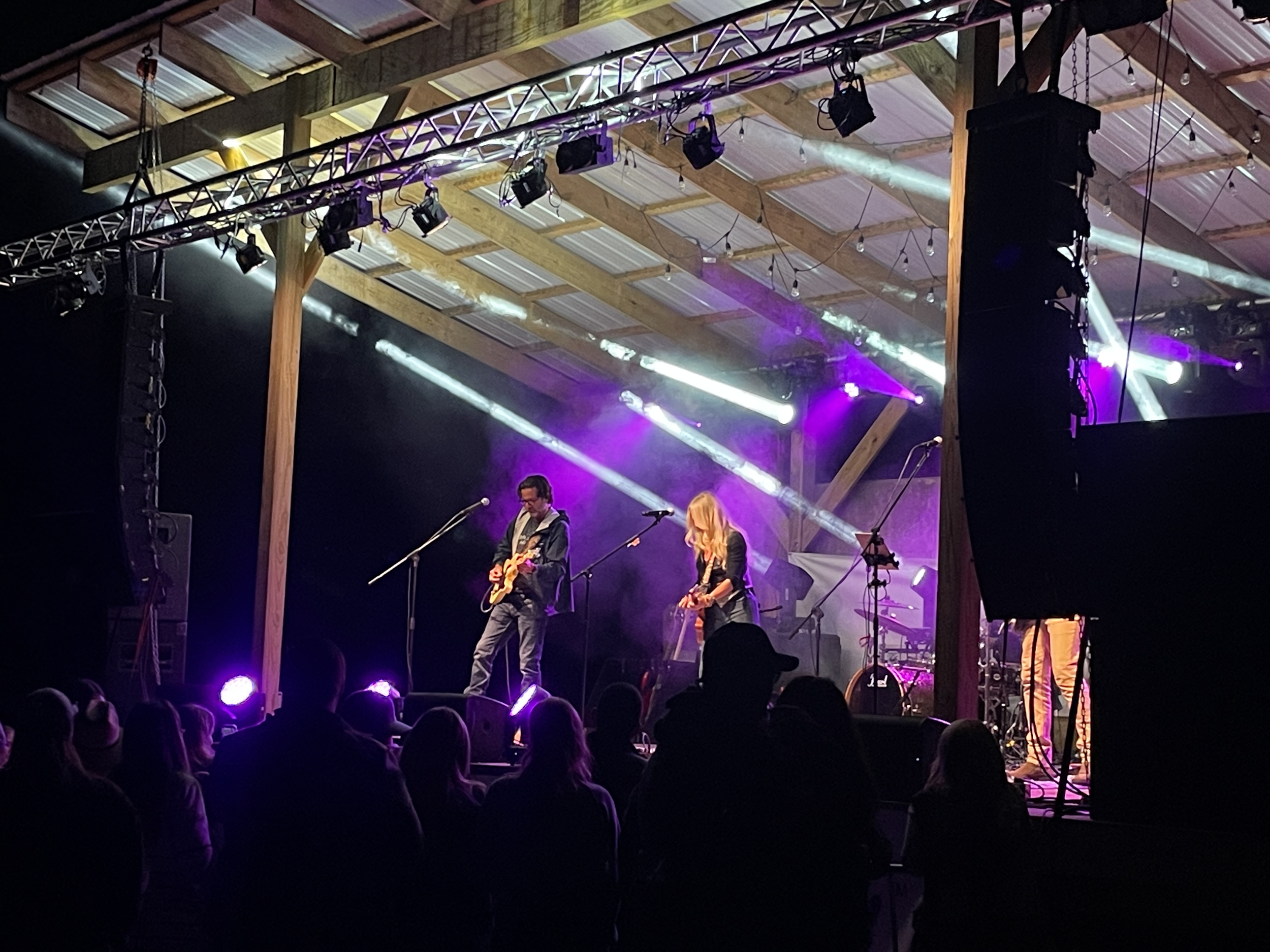 Stage with musicians performing under purple and white lighting, audience silhouettes visible in the foreground, venue appears to be a rustic barn with wood beams and a metal roof.