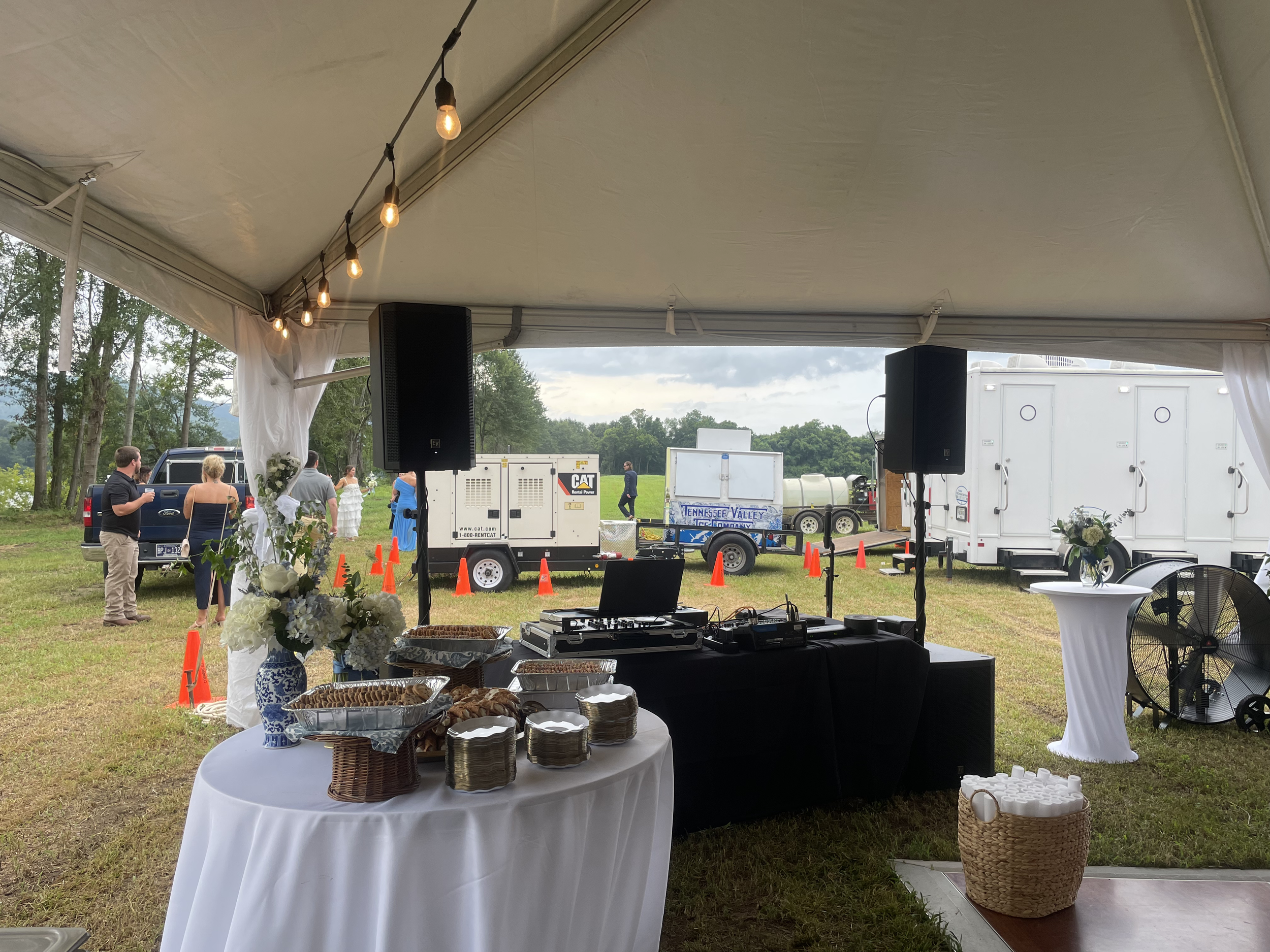 Outdoor event under a large canopy with string lights hanging from the ceiling, serving tables with food, and people socializing in a grassy area surrounded by trees and equipment trailers.