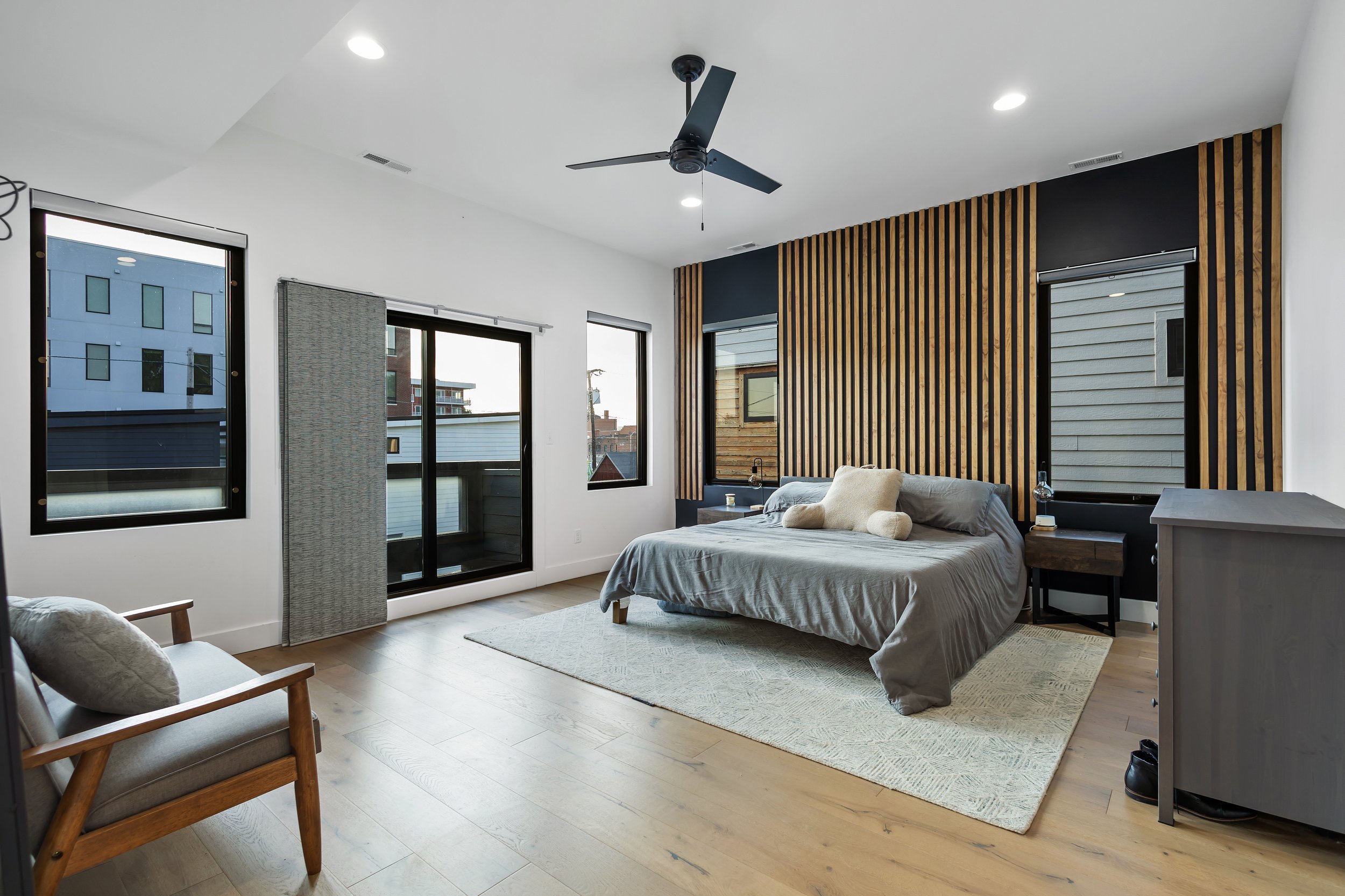 Modern bedroom with wooden feature wall, gray bed with pillows, bedside tables, a gray dresser, and a sitting area with a wooden chair. Large windows and sliding door to balcony, ceiling fan, and recessed lighting.