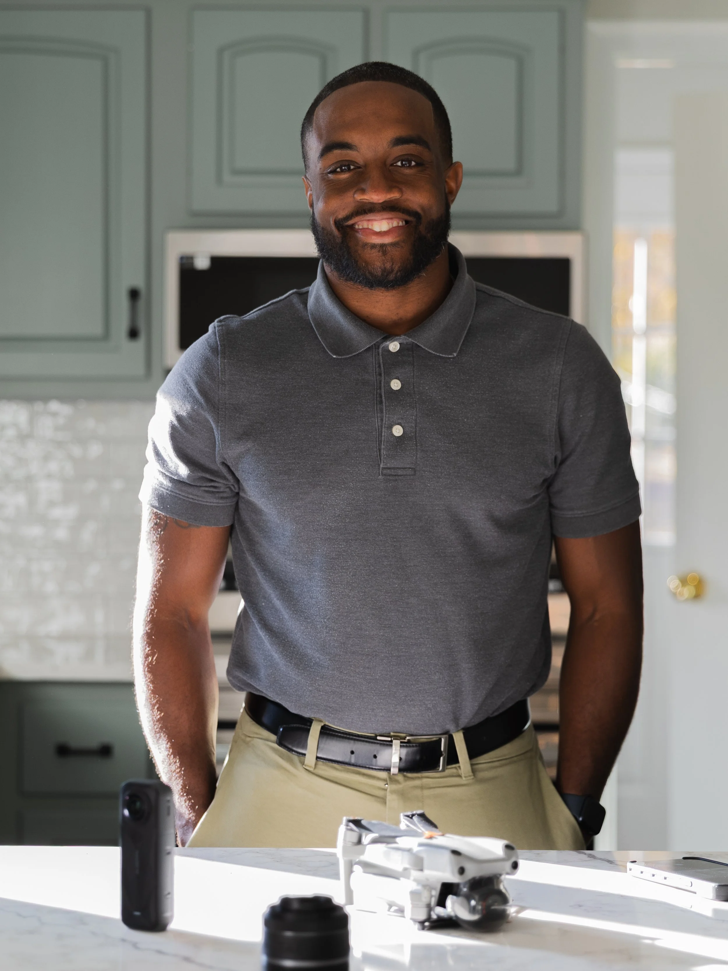A smiling man stands in a kitchen behind a white countertop with a drone, smartphone, and camera lens on it.