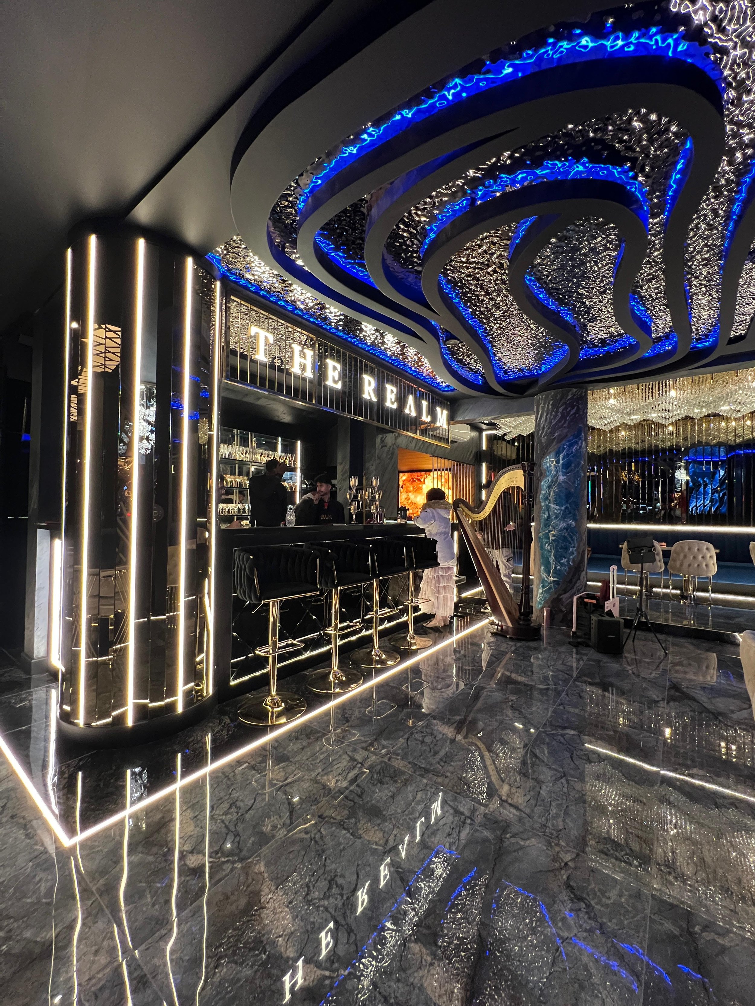Interior of a luxurious bar named 'The Realm' with modern black and gold decor, including a curved bar counter, illuminated signage, reflective black marble flooring, and LED lighting accents.