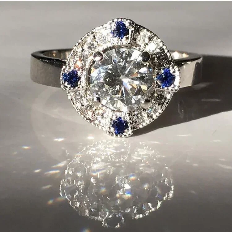 A diamond and sapphire ring with a large central diamond surrounded by smaller diamonds and six blue sapphires, reflecting light and creating a sparkling effect.