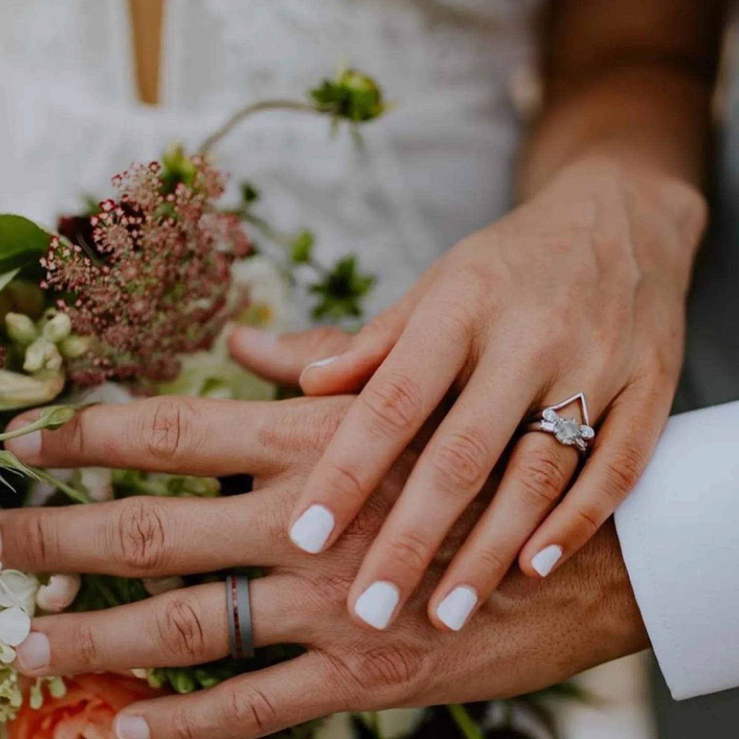Close-up of two hands, one with a diamond engagement ring and the other with a wedding band, resting on a bouquet of flowers.