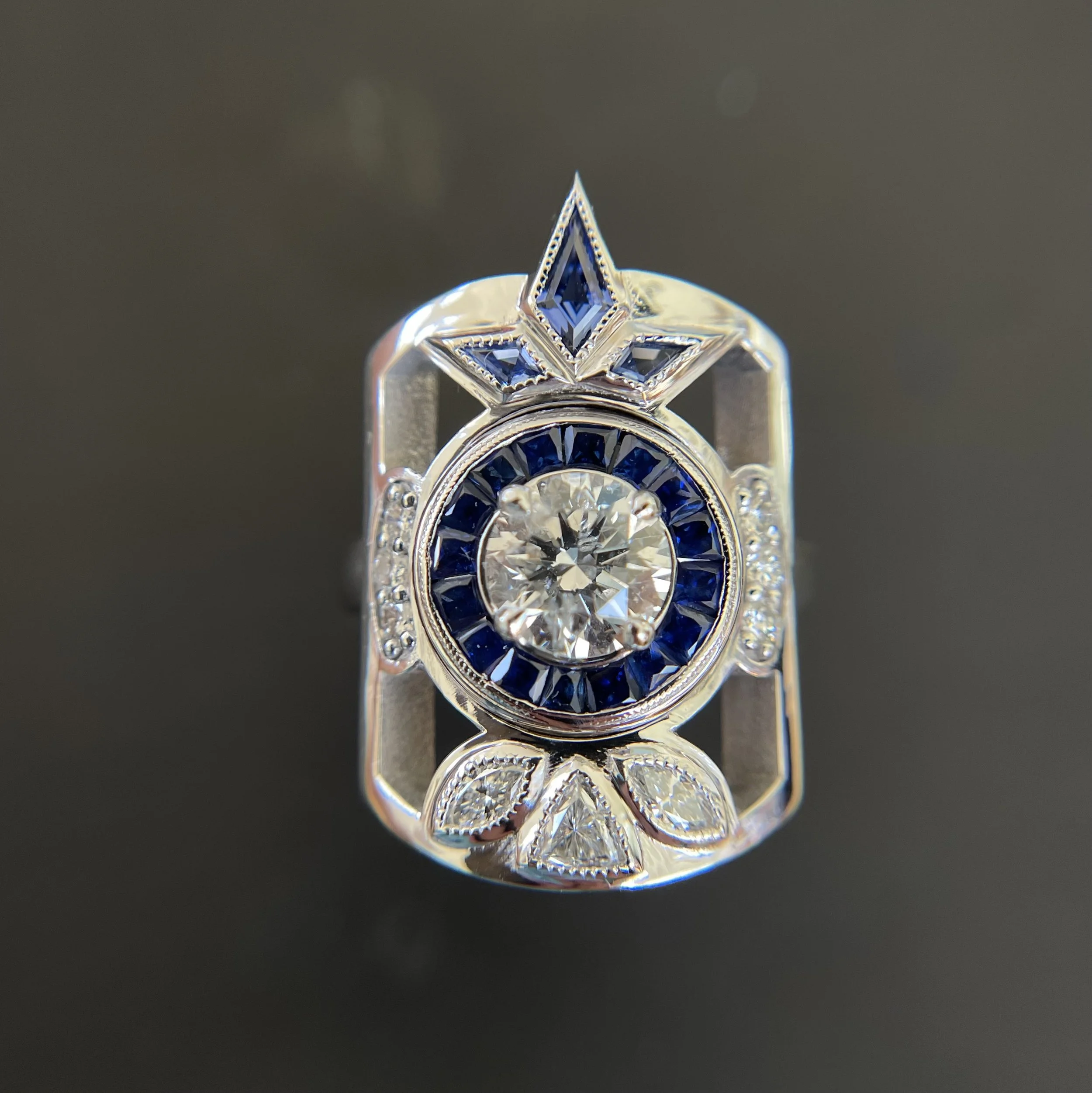 A 14k white gold ring with a large central diamond surrounded by blue sapphires and smaller diamonds, featuring geometric and star-shaped designs.