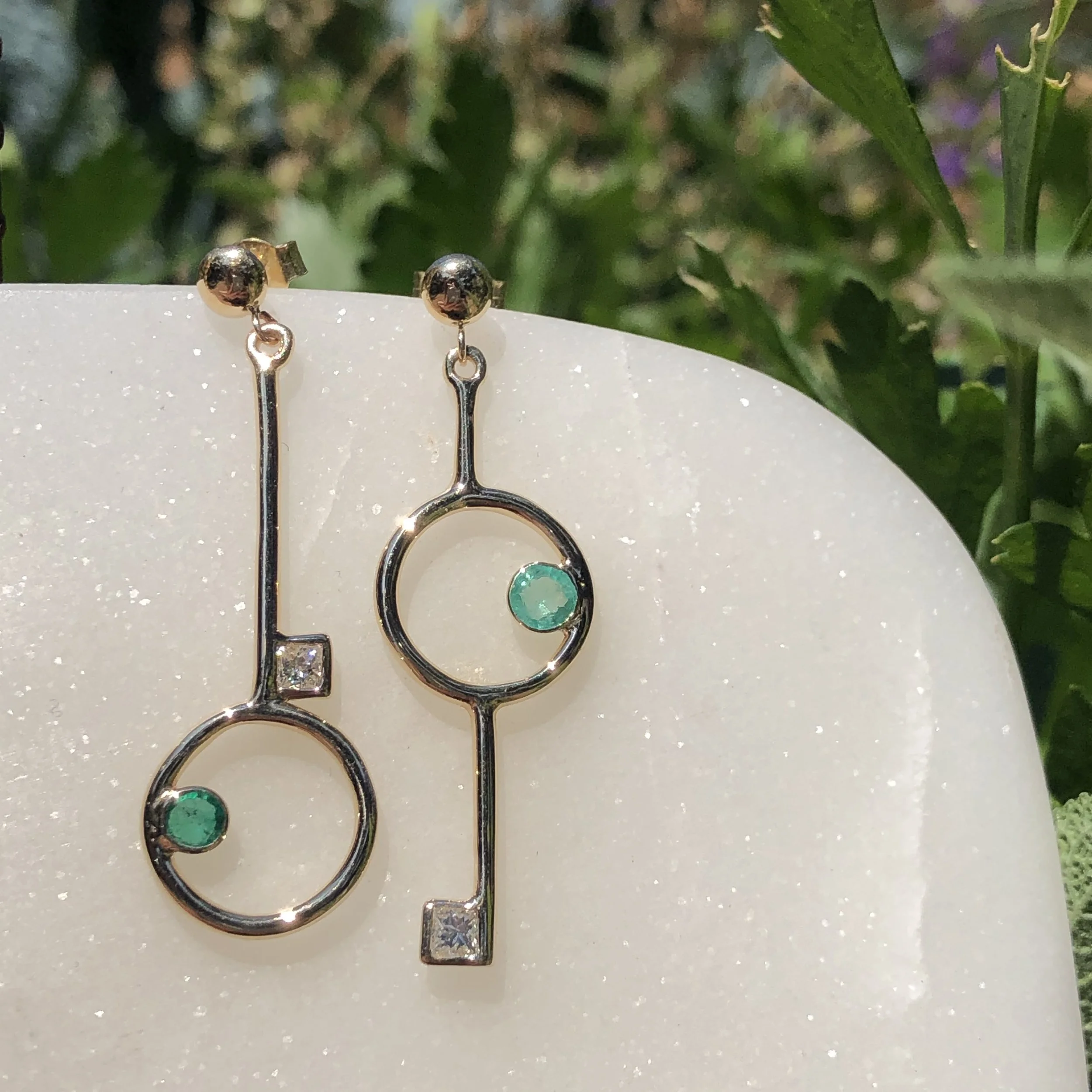 Pair of modern silver earrings with circular and rectangular design, each featuring small green and clear gemstones, displayed on a white sparkly surface outdoors.