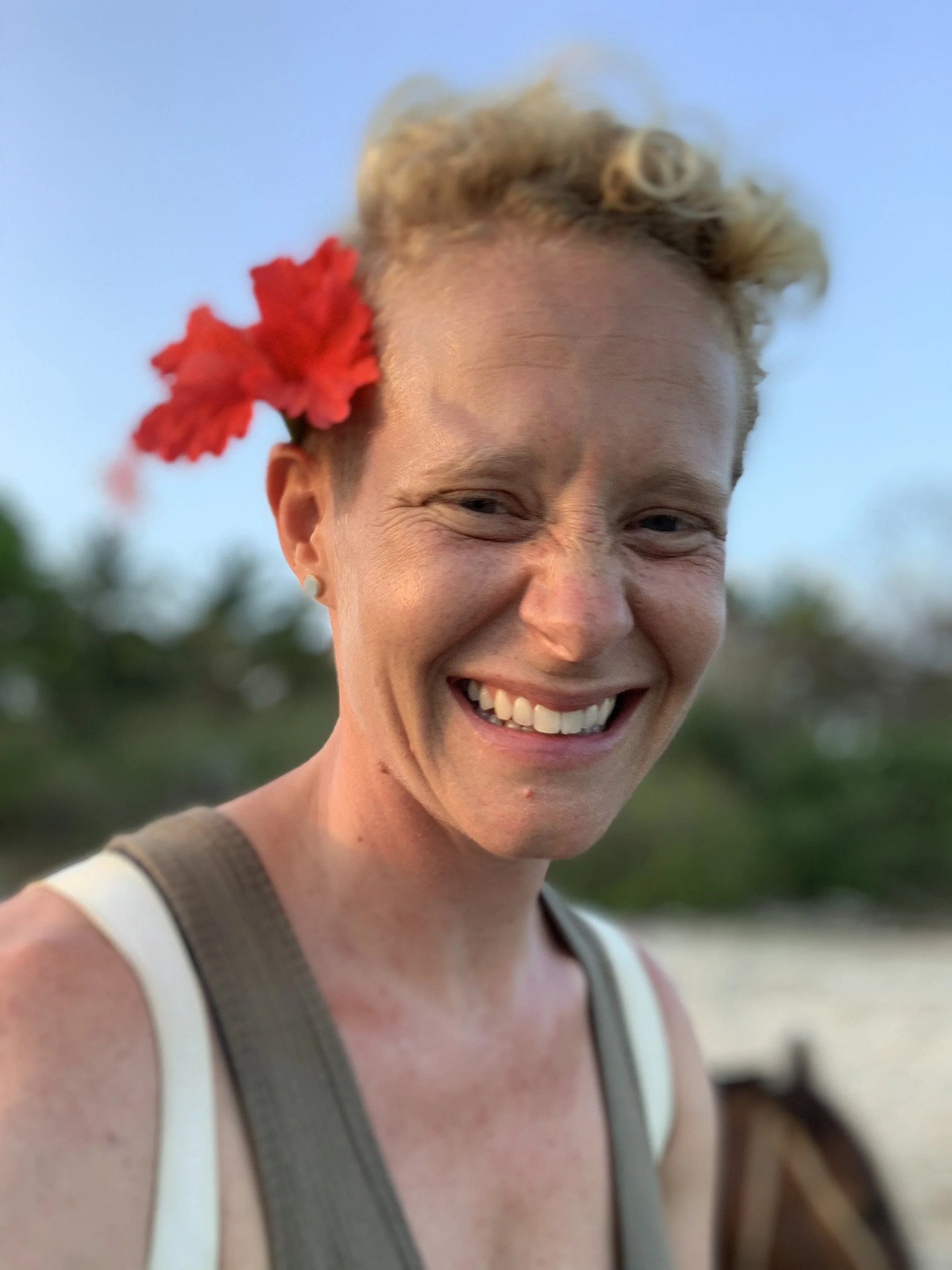A woman with curly blonde hair and a red flower in her hair, smiling outdoors during sunset, wearing a white top with beige straps.