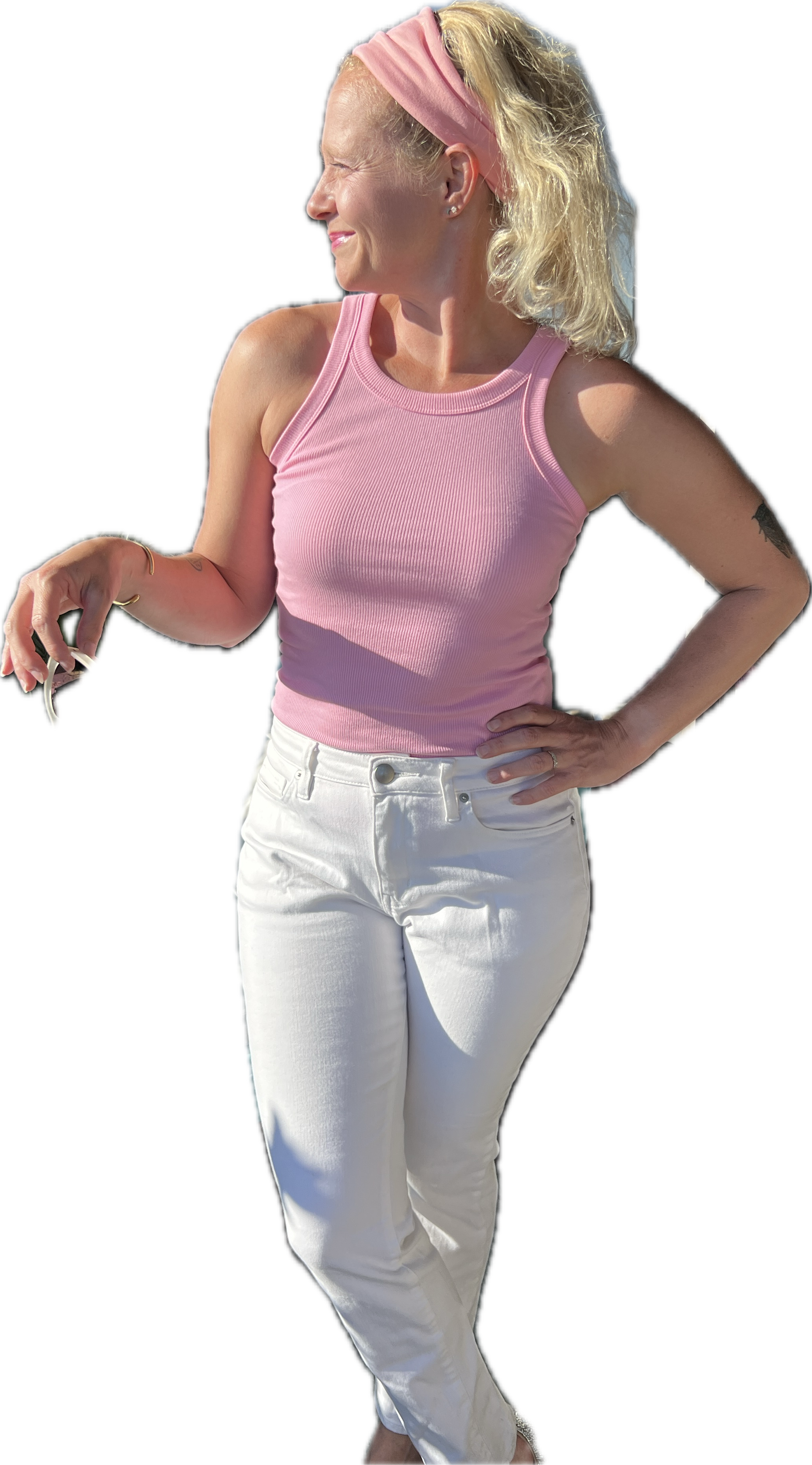 A woman with blonde hair wearing a pink tank top, white pants, a pink headband, and earrings, standing outdoors with her hand on her hip, smiling and looking to the side.